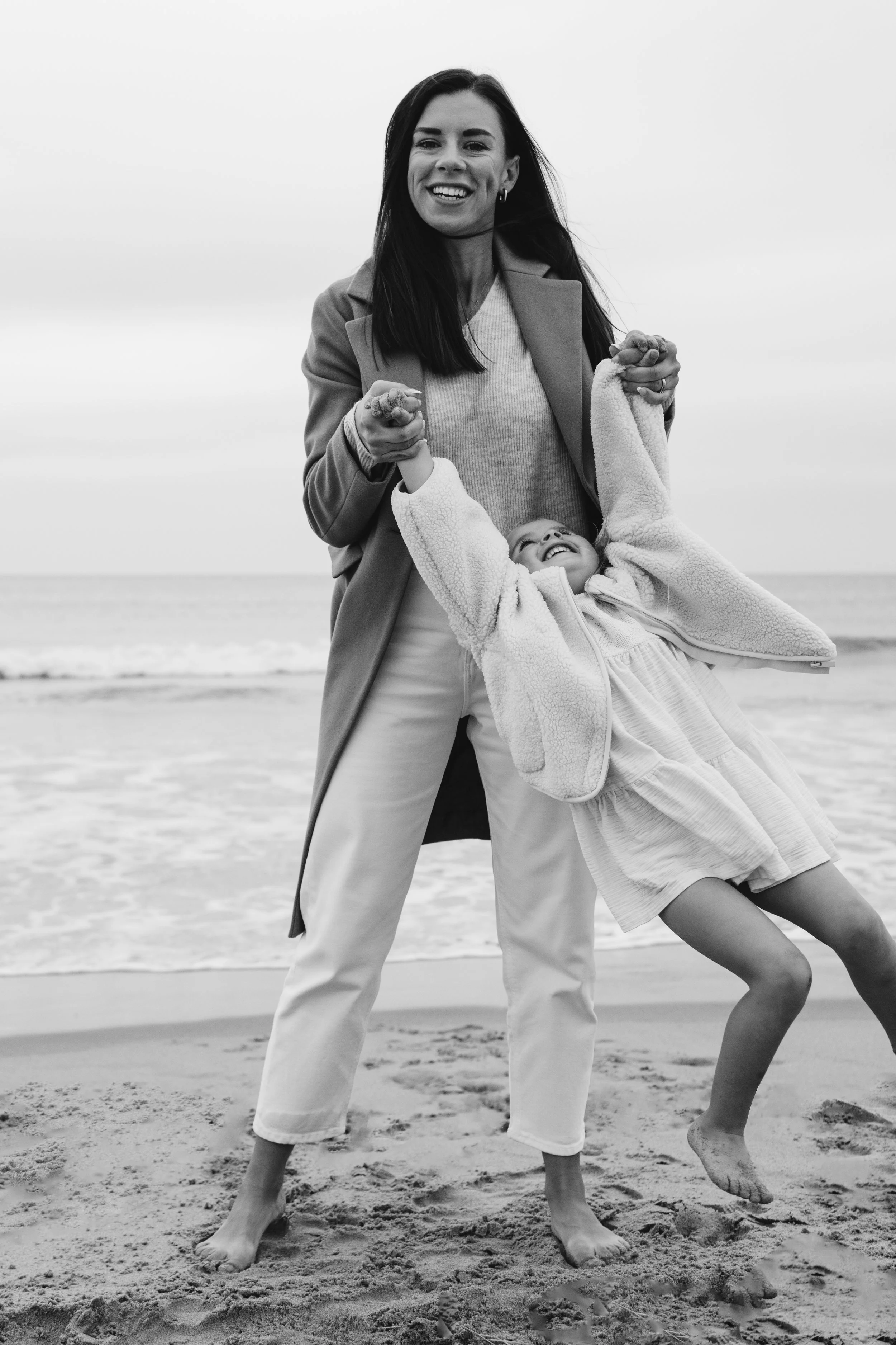 A woman and a child on a beach, holding hands and smiling. The woman is standing while the child is leaning back, both barefoot on the sand, with the ocean in the background.