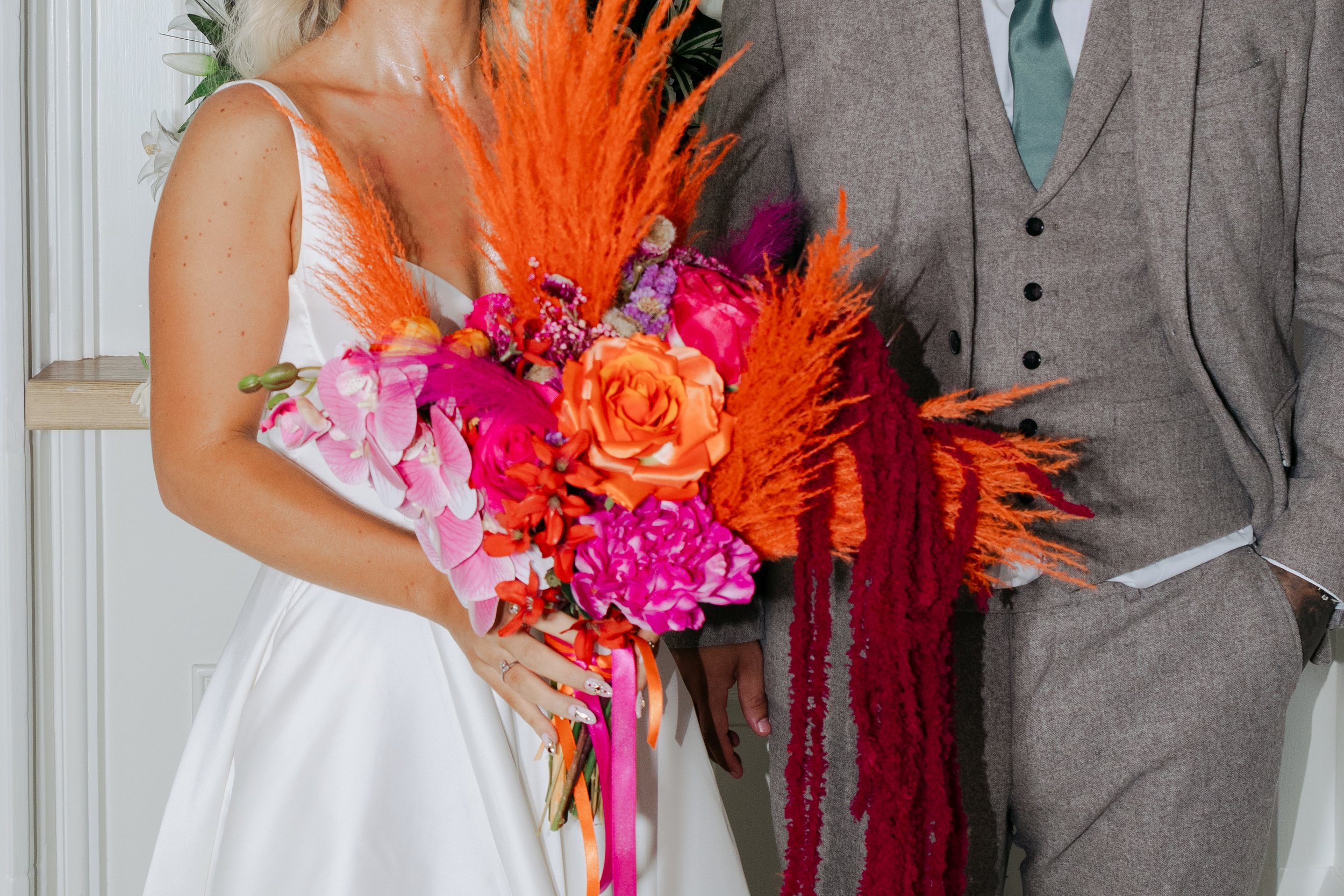 A bride in a white wedding dress holding a vibrant bouquet of pink, orange, purple, and red flowers, with orange and red decorative elements, standing next to a groom in a gray suit.