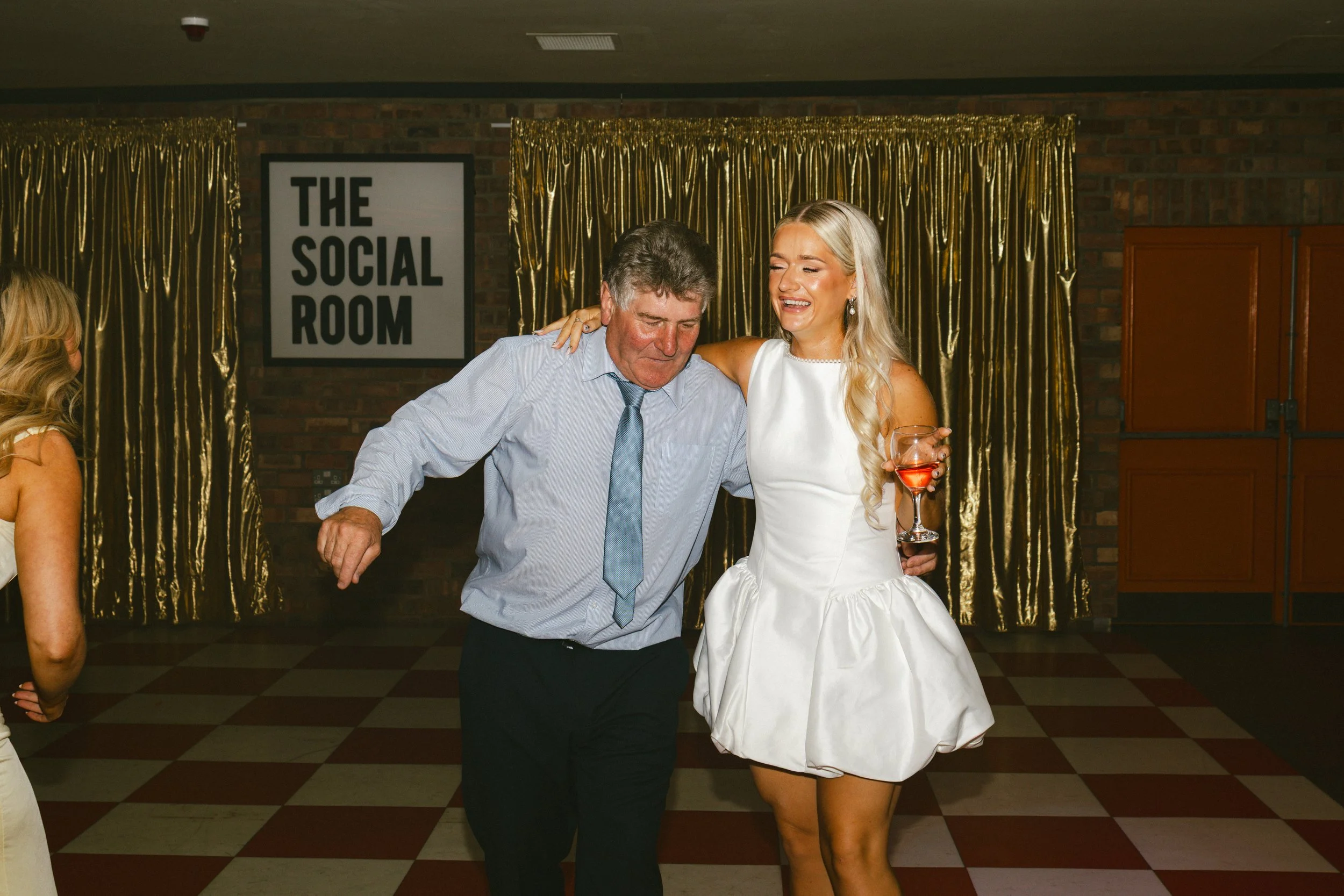 A man and woman dancing together at a party, with the woman holding a drink, in front of a gold curtain backdrop and a sign that reads 'The Social Room'.
