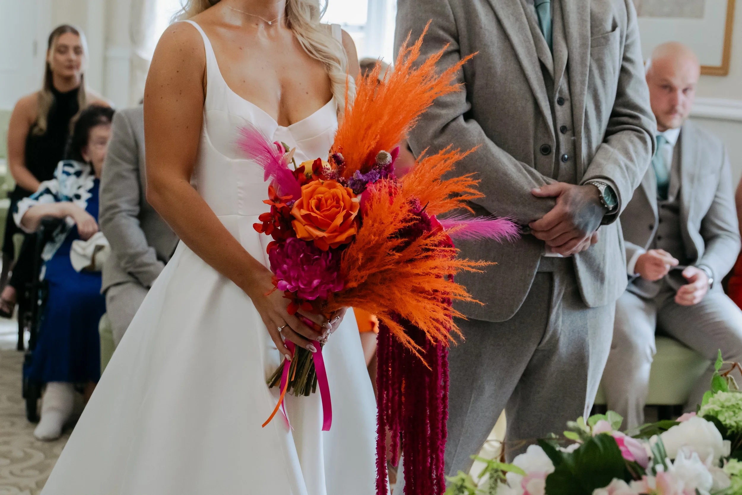 A woman in a white dress holding a colorful bouquet of flowers including orange roses, pink feathers, and orange and pink pampas grass during a wedding ceremony.
