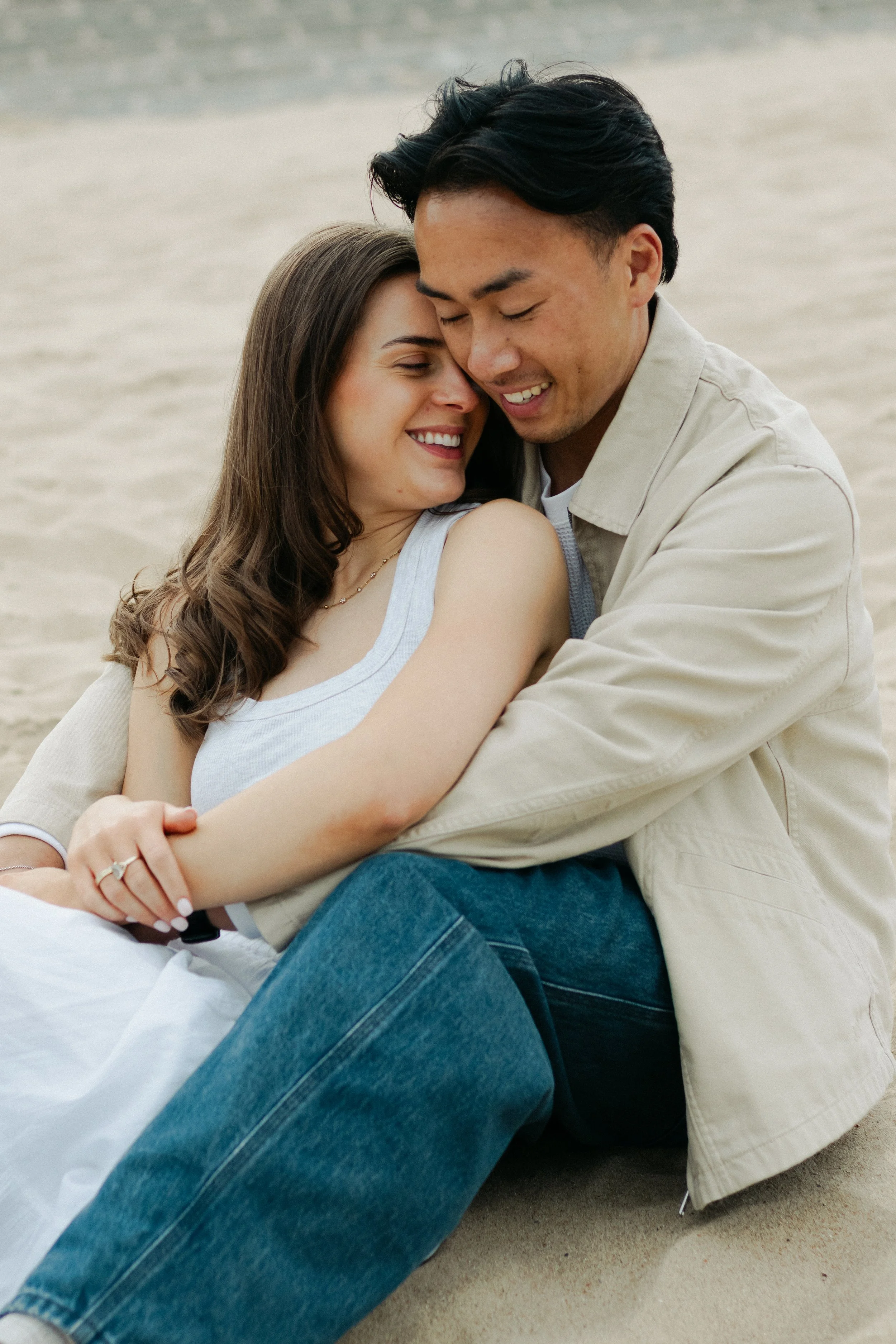 A smiling couple hugging on the beach, with the woman in a white tank top and the man in a beige jacket, both appearing happy and relaxed.
