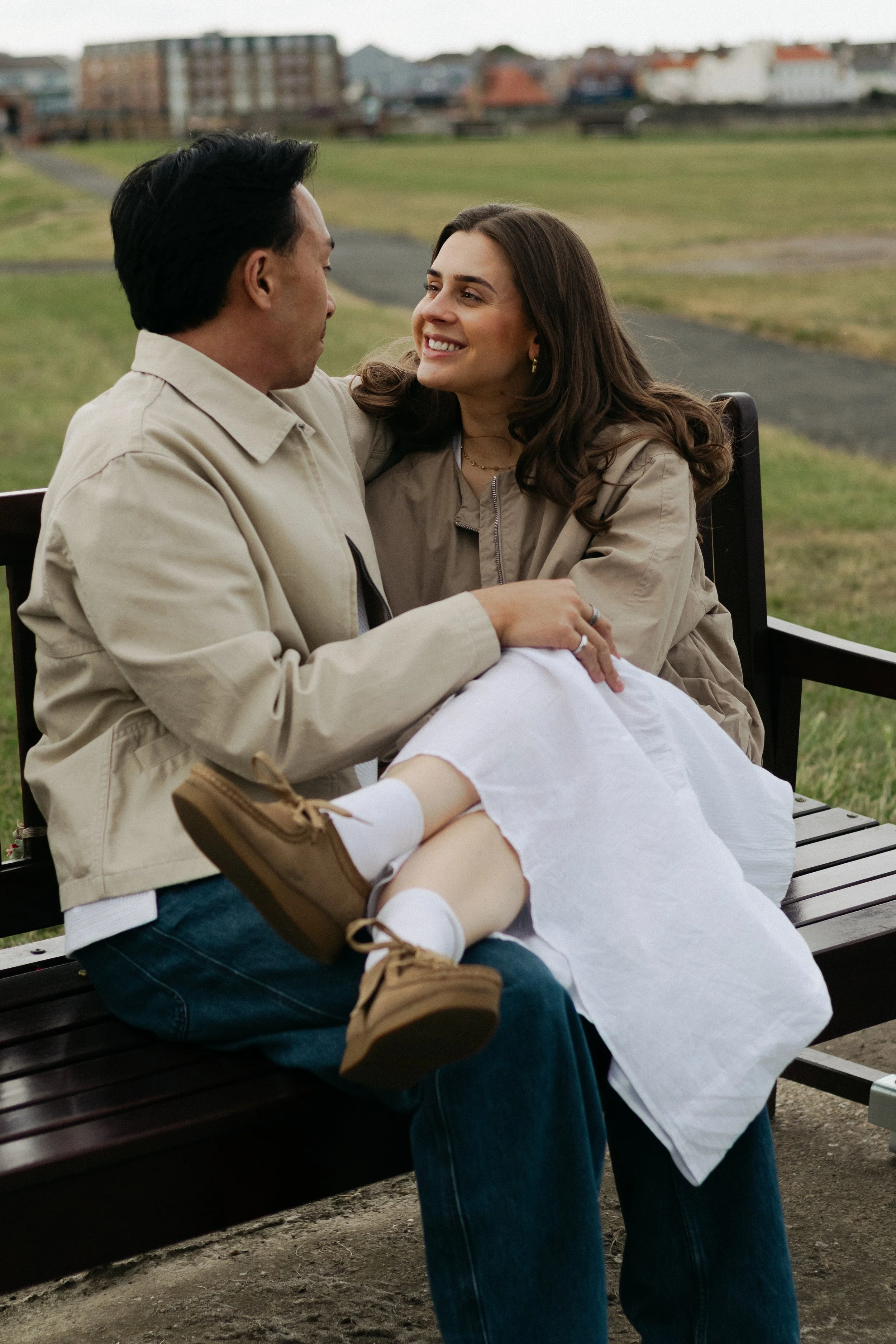 A young woman with long brown hair smiling at a young man with dark hair, sitting on a park bench outdoors. The woman has her arm around the man's shoulders, and they are looking at each other. The background features a grassy park and buildings in the distance.