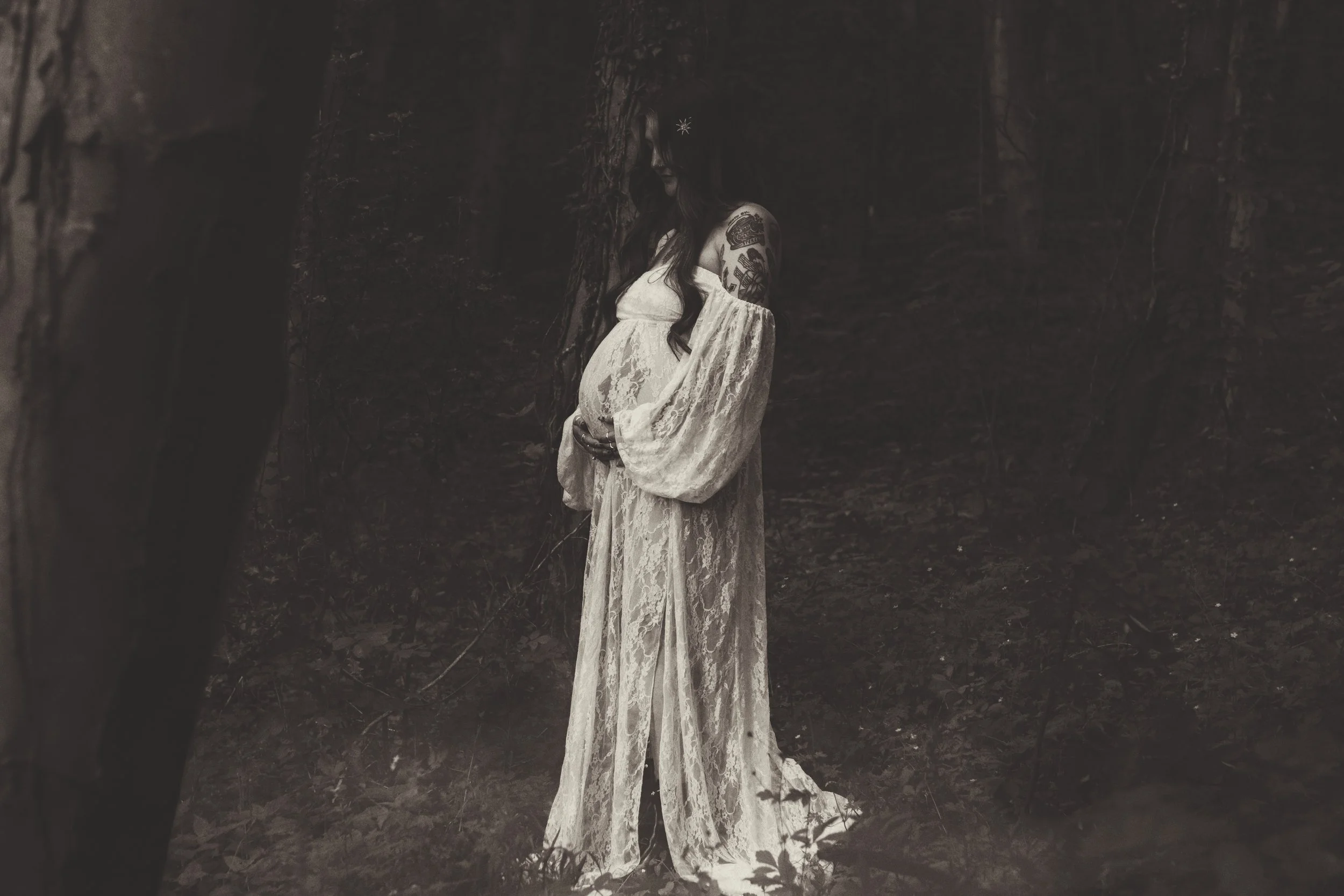 A pregnant woman in a white lace dress standing in a dark forest with trees and leaves visible around her, holding her belly and looking down.