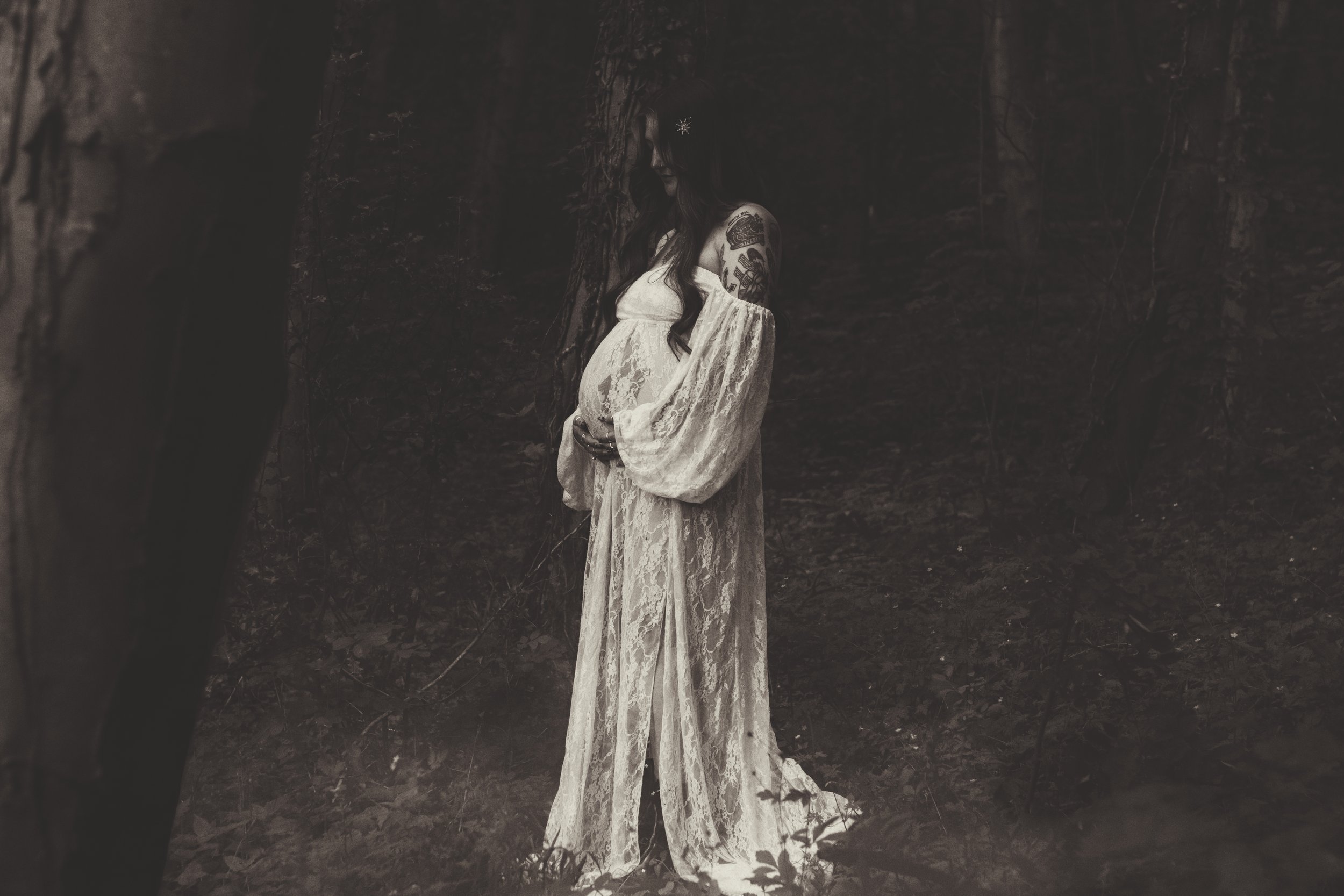 A pregnant woman in a long, lace dress standing in a dark forest with trees and leaves surrounding her in warm.