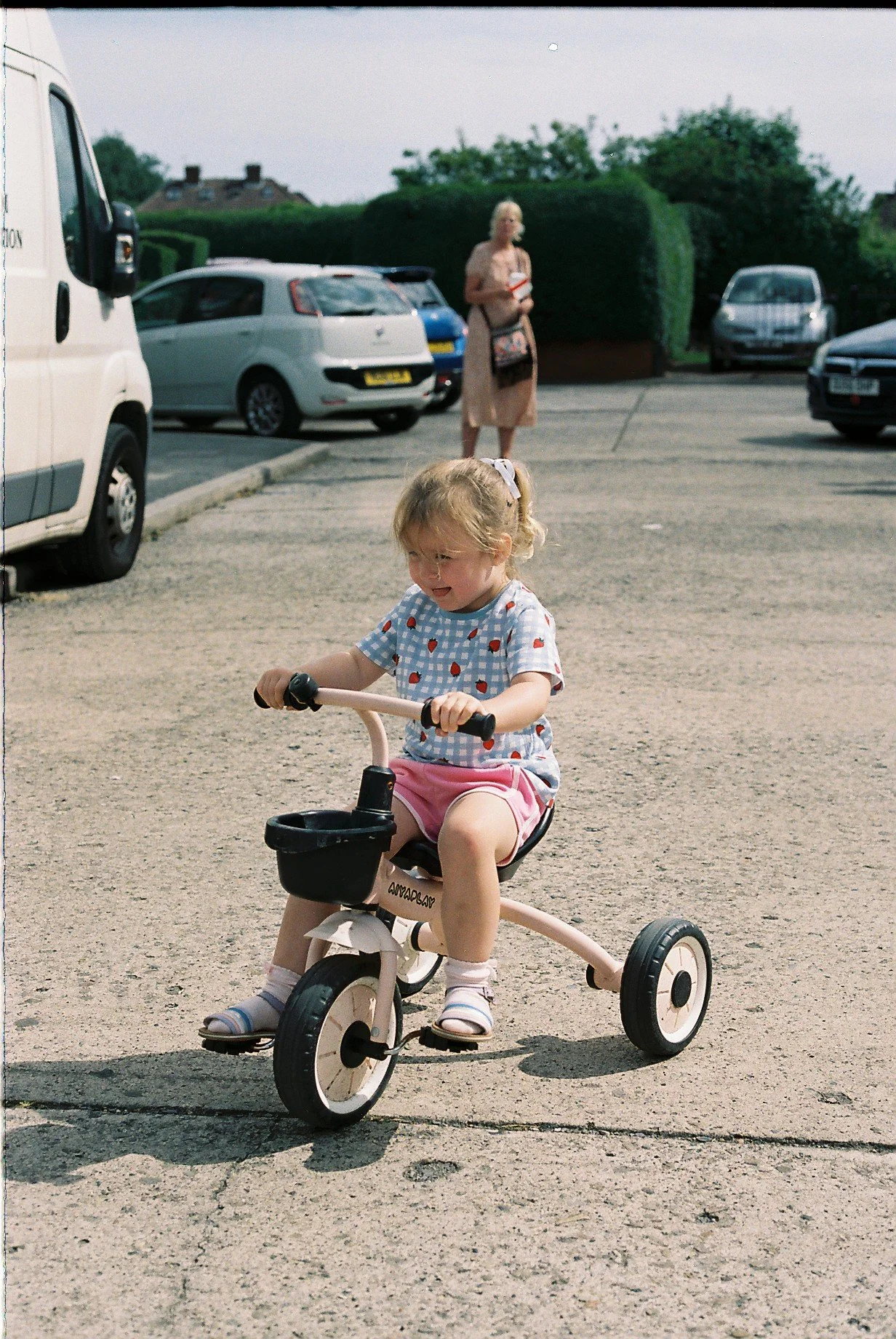 A young girl riding a small pink pedal tricycle with a black basket in a parking lot. She is wearing a blue checkered shirt with red strawberry patterns, pink shorts, and sandals. In the background, there is a woman standing near parked cars and large green bushes.