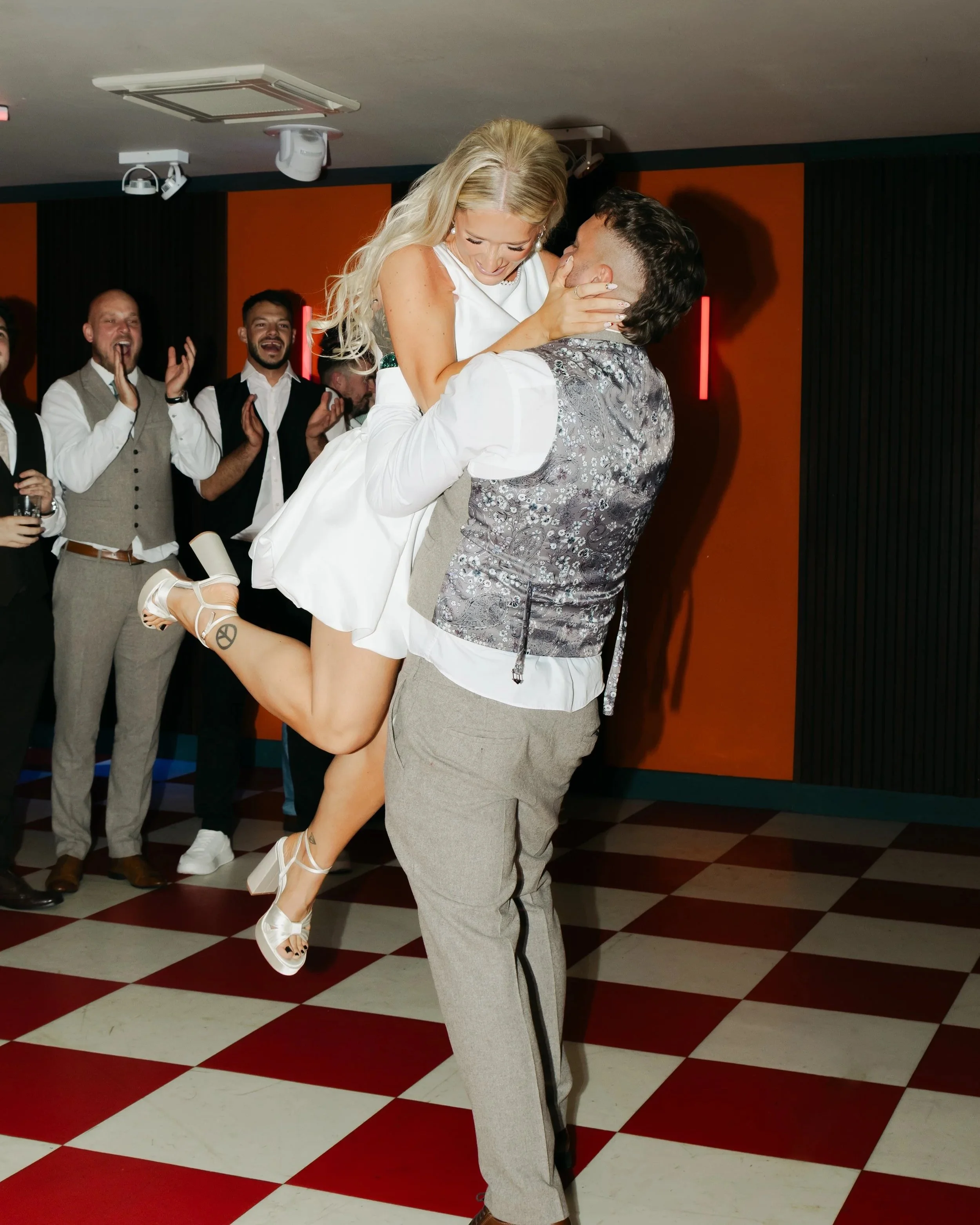 A man lifts a woman in a white dress during a celebration or party, with people clapping and cheering in the background.