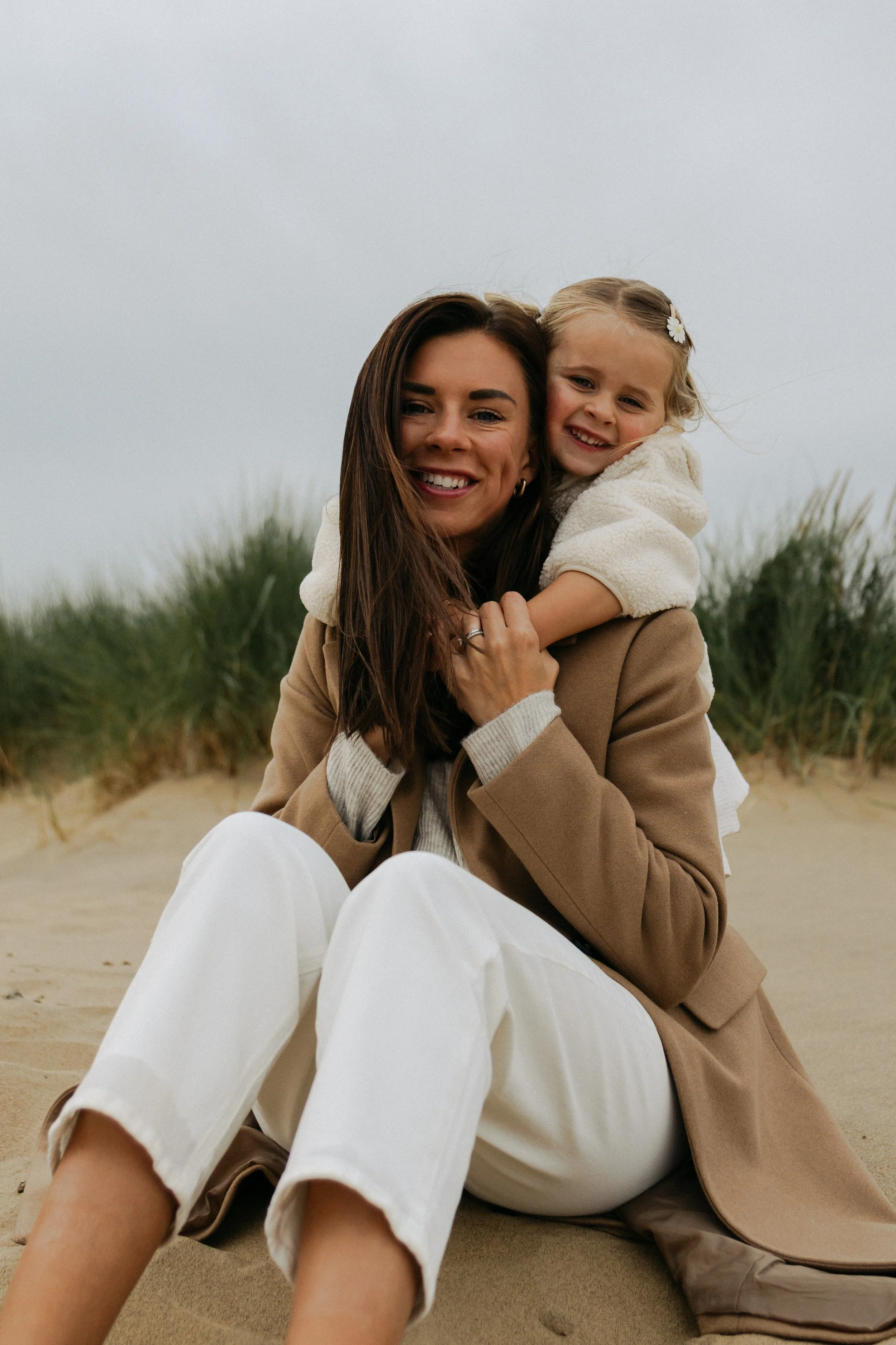 A woman with long brown hair and a young girl with blonde hair and a white flower hair clip embrace on a beach, smiling at the camera.