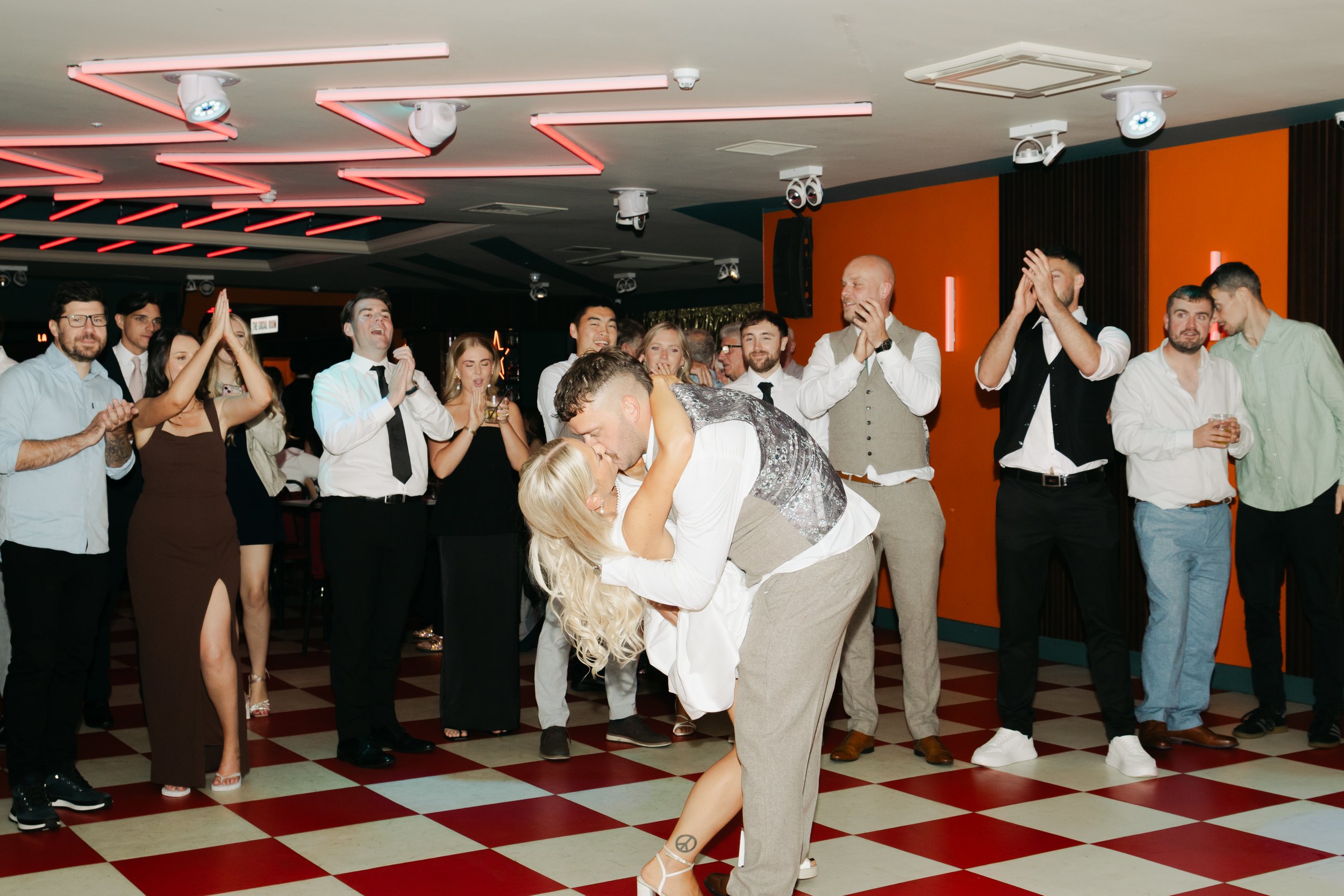 A group of people celebrating at a party, with a couple dancing in the center, surrounded by friends clapping and smiling, in a room with orange and black walls and checkered floor.