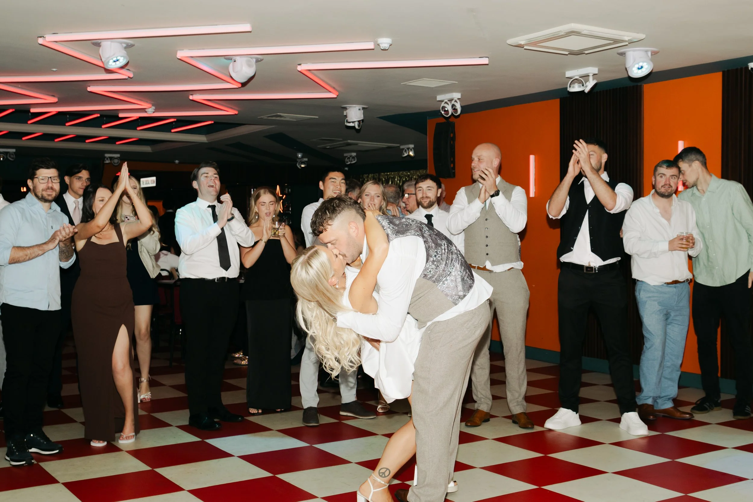 A group of people celebrating at a party, with a couple dancing in the center, surrounded by friends clapping and smiling, in a room with orange and black walls and checkered floor.