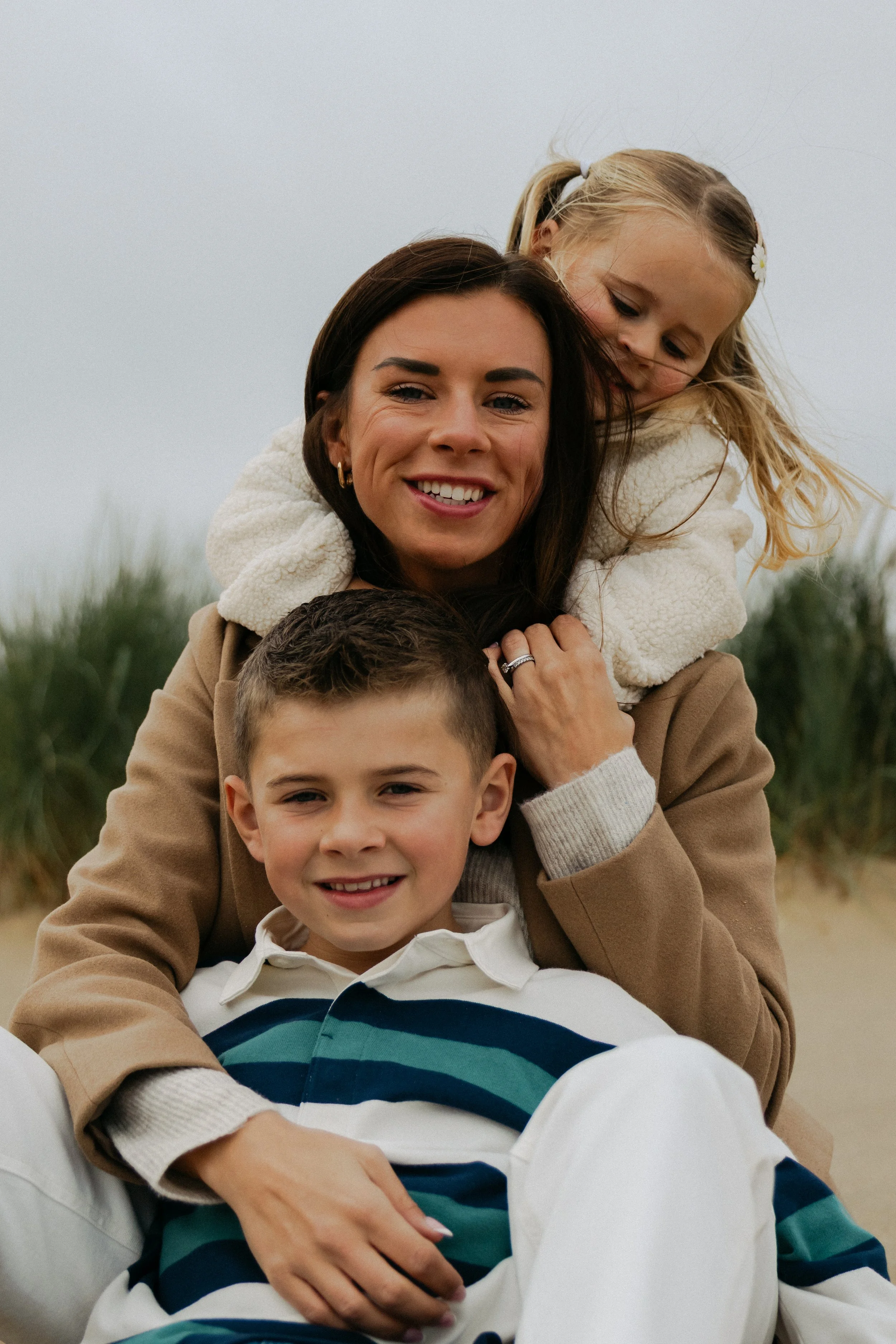 A woman with dark hair smiling, a young girl with blonde hair and a white hair clip hugging her from behind, and a young boy with short hair and a striped sweater sitting in front, outdoors with a cloudy sky and greenery in the background.