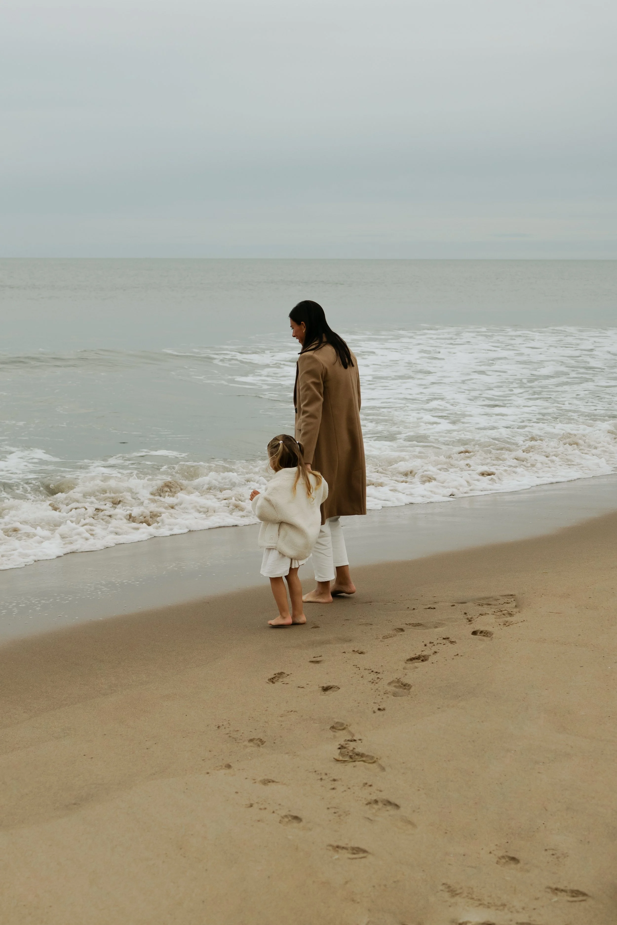 A woman and a young girl walking along the beach, leaving footprints in the sand, with the ocean waves nearby and a cloudy sky overhead.