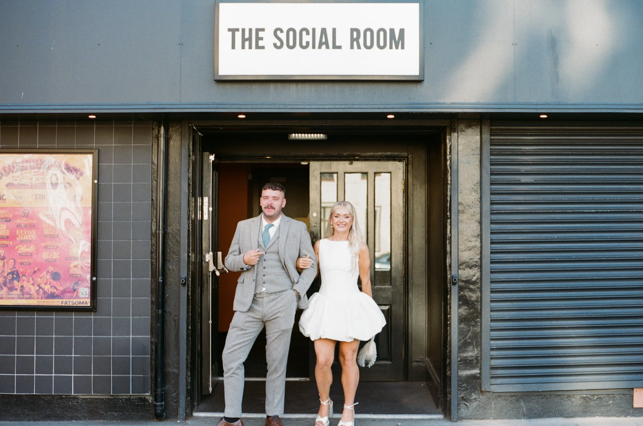 A man and a woman dressed in formal attire standing in front of a building labeled "The Social Room." The man is wearing a gray suit with a blue tie and is pointing to his chest, while the woman is in a white dress with lace details and heels, smiling at the camera.