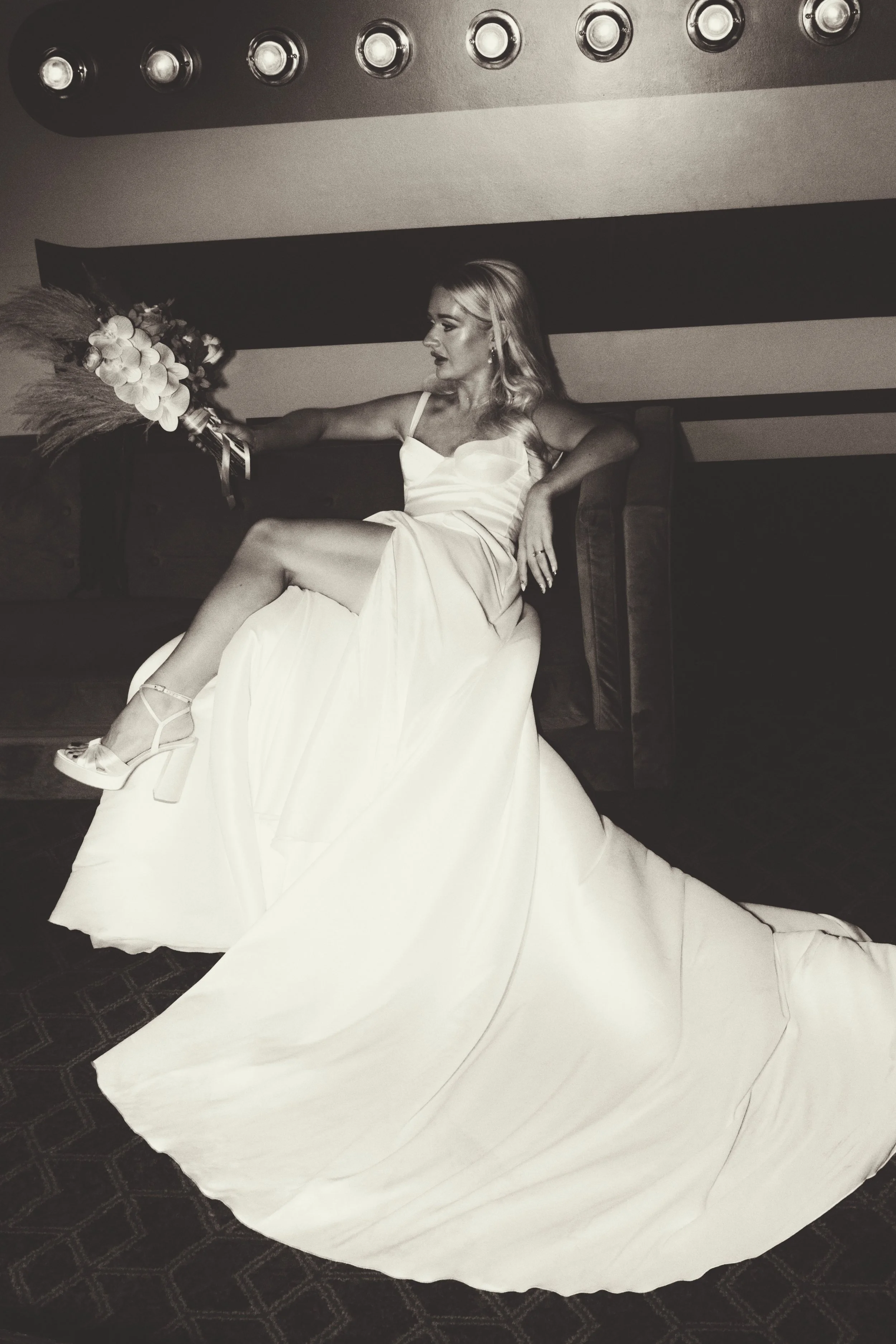 A woman in a wedding dress sitting on a chair, holding a bouquet of flowers, with a dark striped background and a patterned carpet.