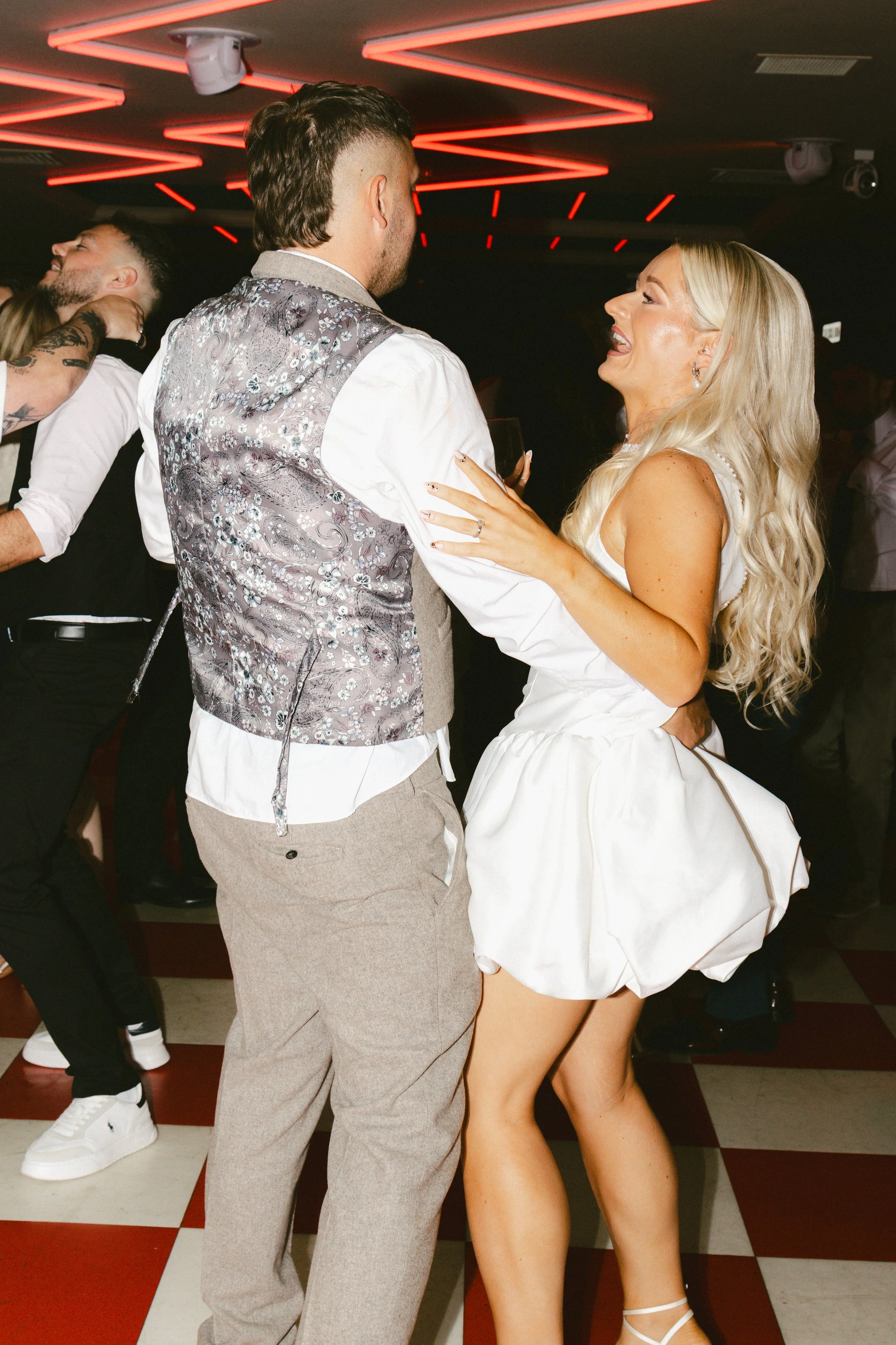 A woman in a white dress dancing with a man in a gray vest at a party or nightclub, with others dancing in the background under red neon lighting.