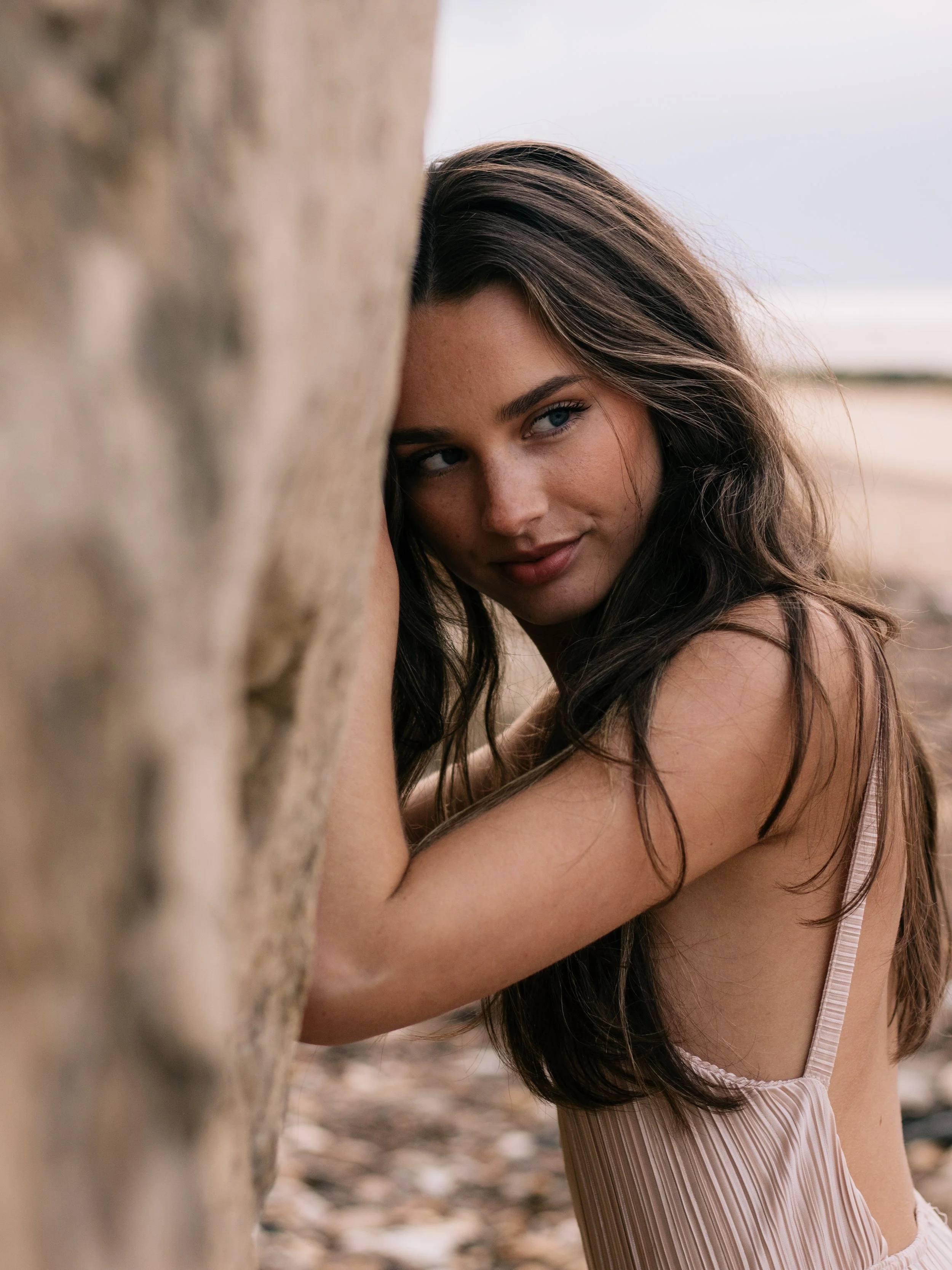 A woman with wavy brown hair and blue eyes peeking out from behind a large rock or tree in a natural outdoor setting with a blurred background.