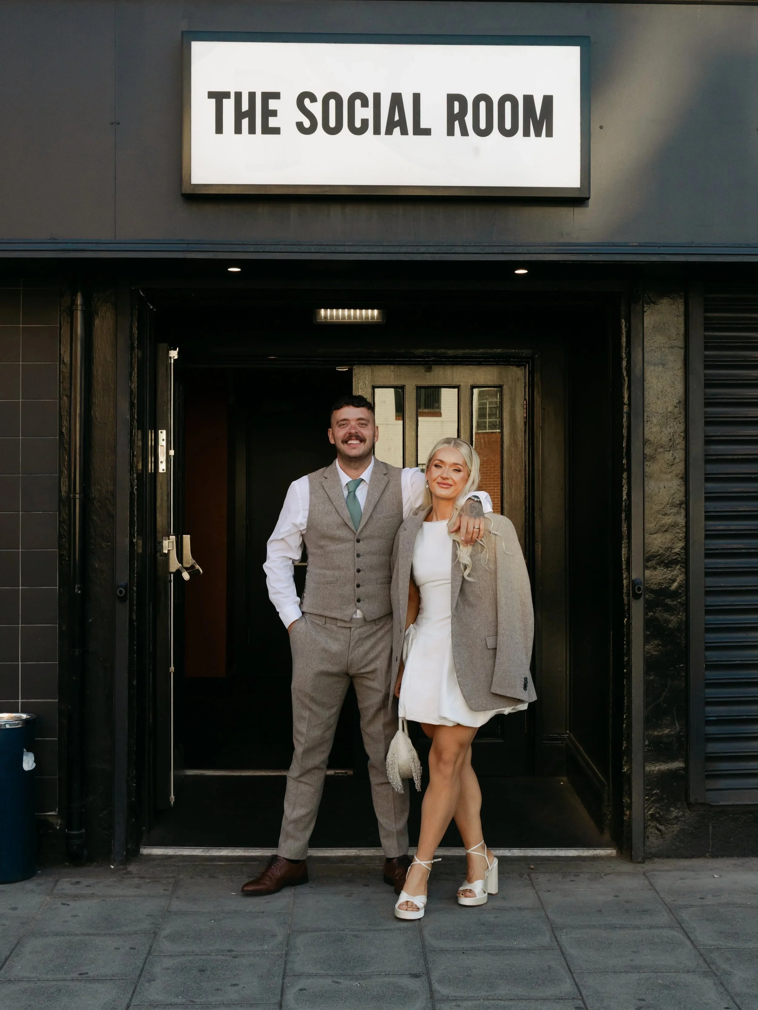 A couple dressed in formal attire standing outside a building with a sign that reads 'The Social Room.' The man is wearing a gray vest, light blue shirt, and tie, with his arm around the woman. The woman is wearing a white dress, gray blazer, and white high-heeled shoes, holding a small purse and smiling.