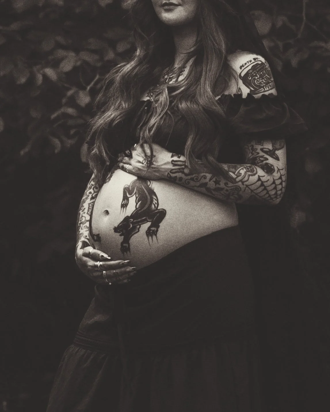 A pregnant woman with long wavy hair, covered in tattoos, standing outdoors in front of leafy background, holding her belly with one hand.