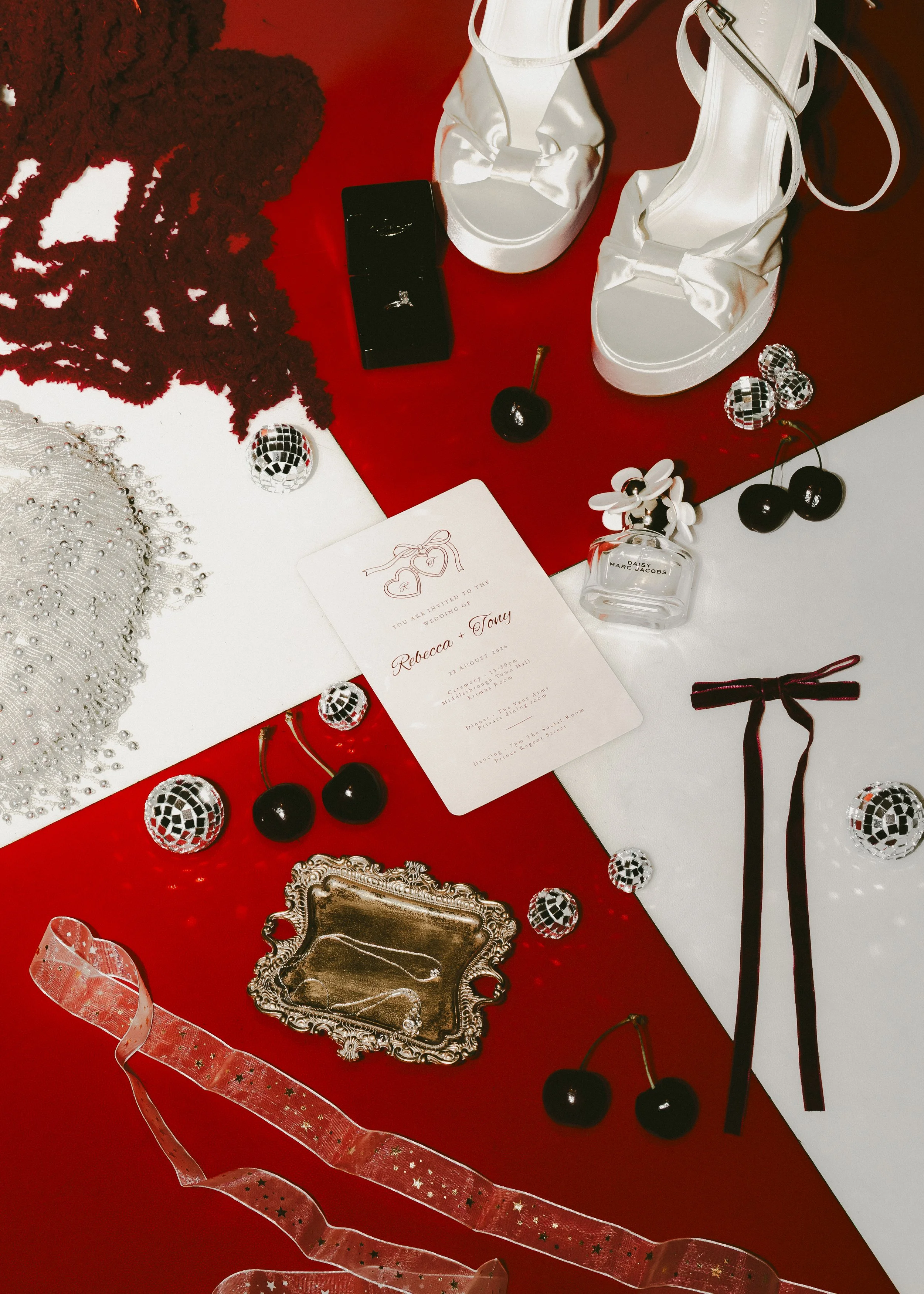 A collection of decorative and jewelry items arranged on a table, including white satin high-heeled shoes with bows, a perfume bottle labeled 'Daisy Marc Jacobs', black and red cherries, jewelry such as earrings and rings, a silver ornate tray, a silver lace fabric, a black jewelry box with a ring, a pink ribbon with star patterns, a red tablecloth, and a white wedding invitation with red and black text.