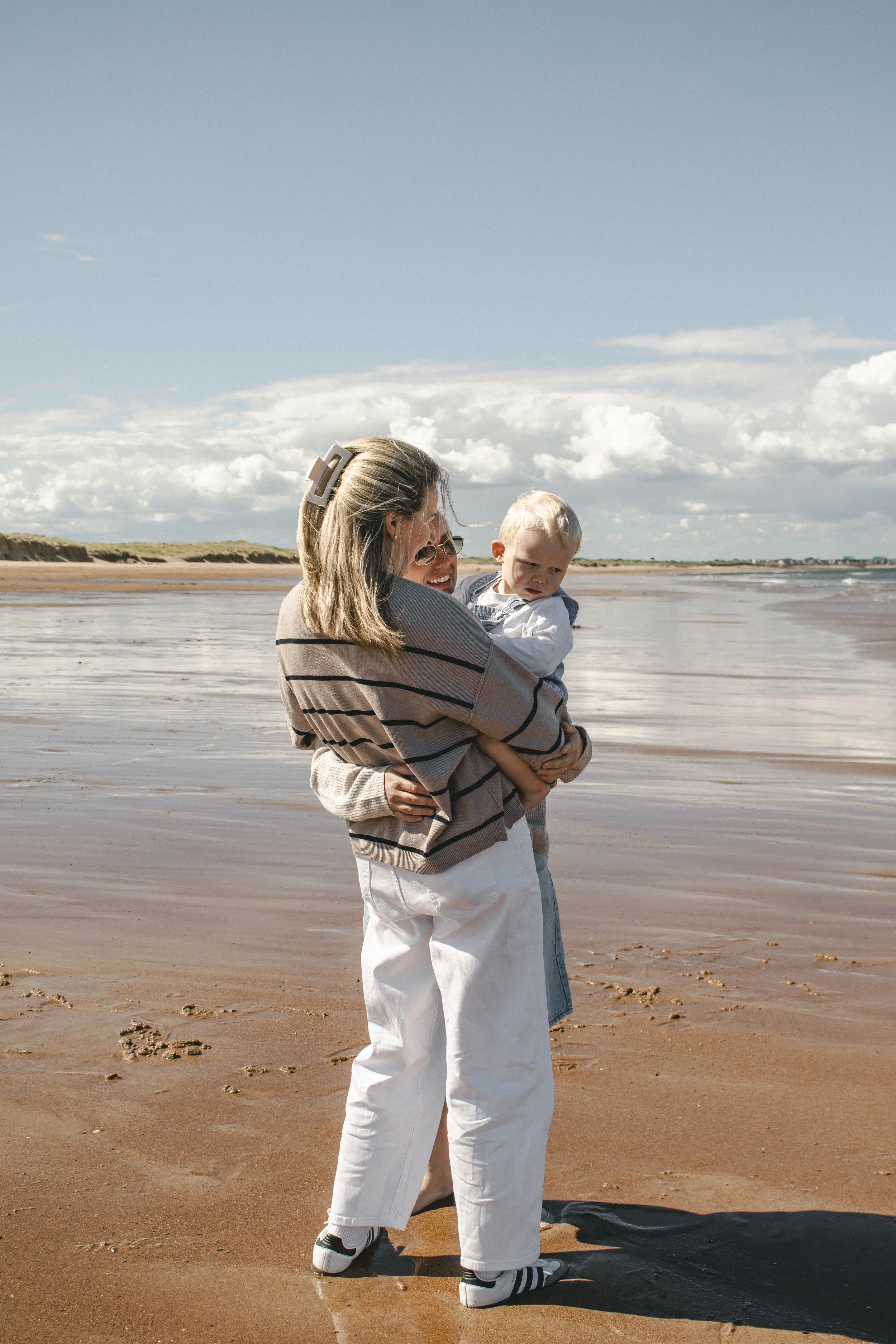 A woman with glasses and a sunhat holding a young boy on the beach, both smiling, with clouds and sea in the background.