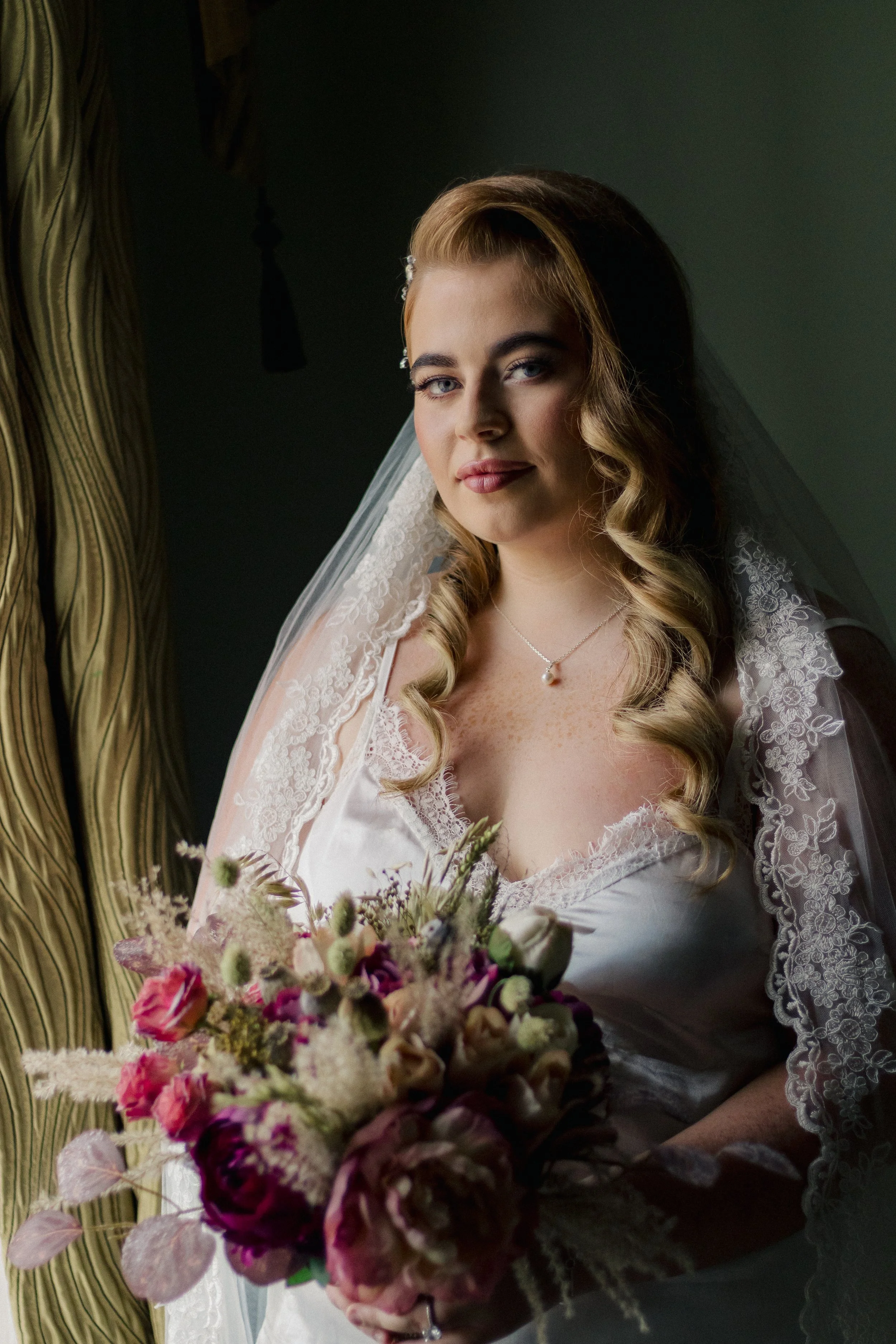 A woman with light skin and blonde, loosely curled hair wears a white lace wedding dress and veil, holding a bouquet of pink, purple, and white flowers while standing indoors near a curtain.