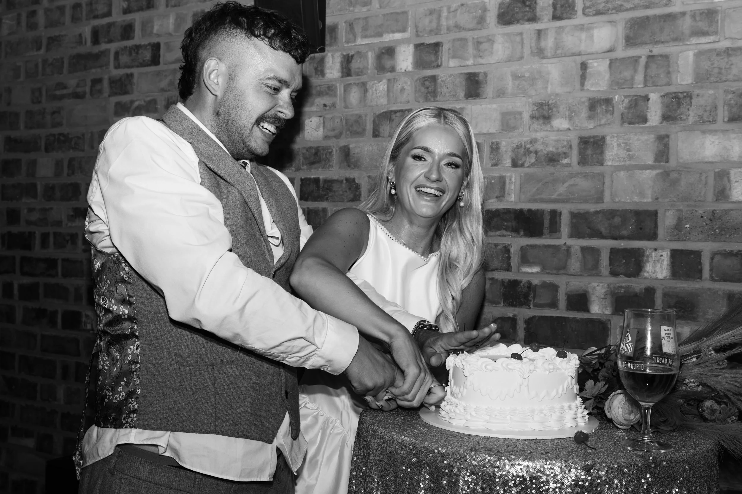 A man and woman are cutting a wedding cake together at a celebration. The man has a beard and curly hair, and is smiling as he looks at the cake. The woman has long blonde hair, earrings, and is also smiling as they cut the cake. A brick wall is in the background, and a drink is on the table nearby.
