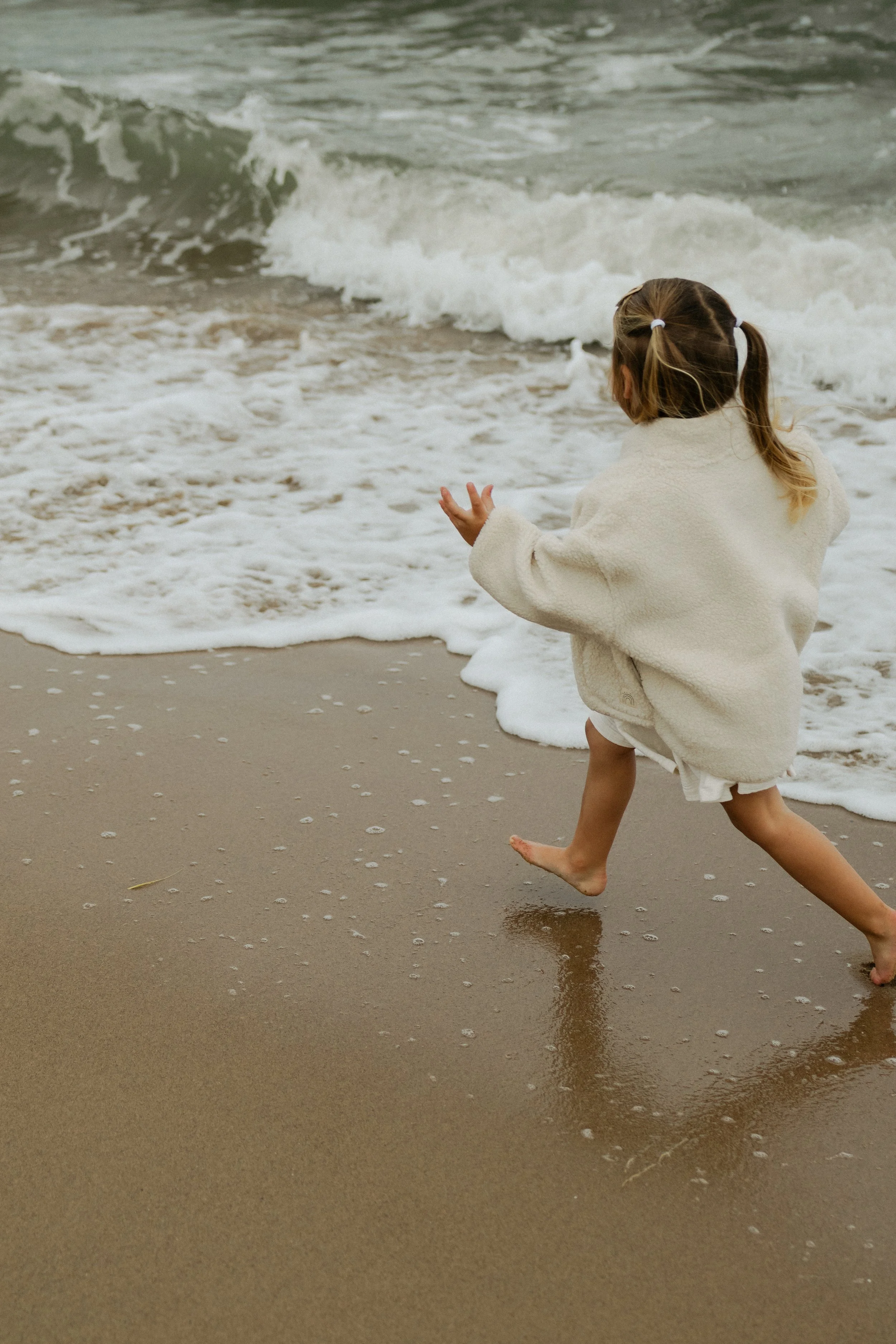 A young girl with two ponytails running barefoot along the shoreline at the beach, with waves crashing onto the sand in the background.