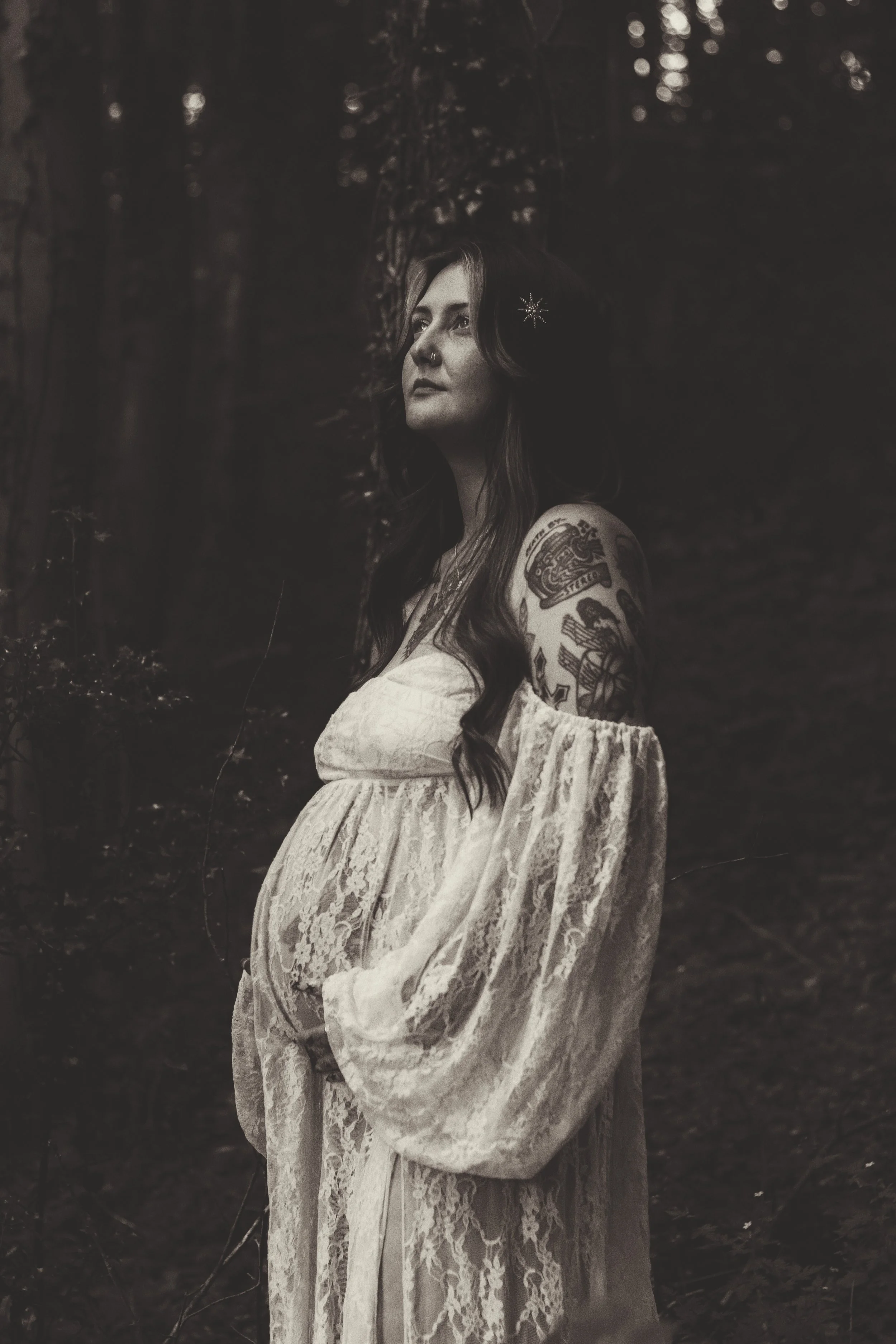 Black and white portrait of a tattooed woman with long dark hair standing outdoors in a forest. She wears a white lace off-shoulder dress and has a star-shaped hair accessory.