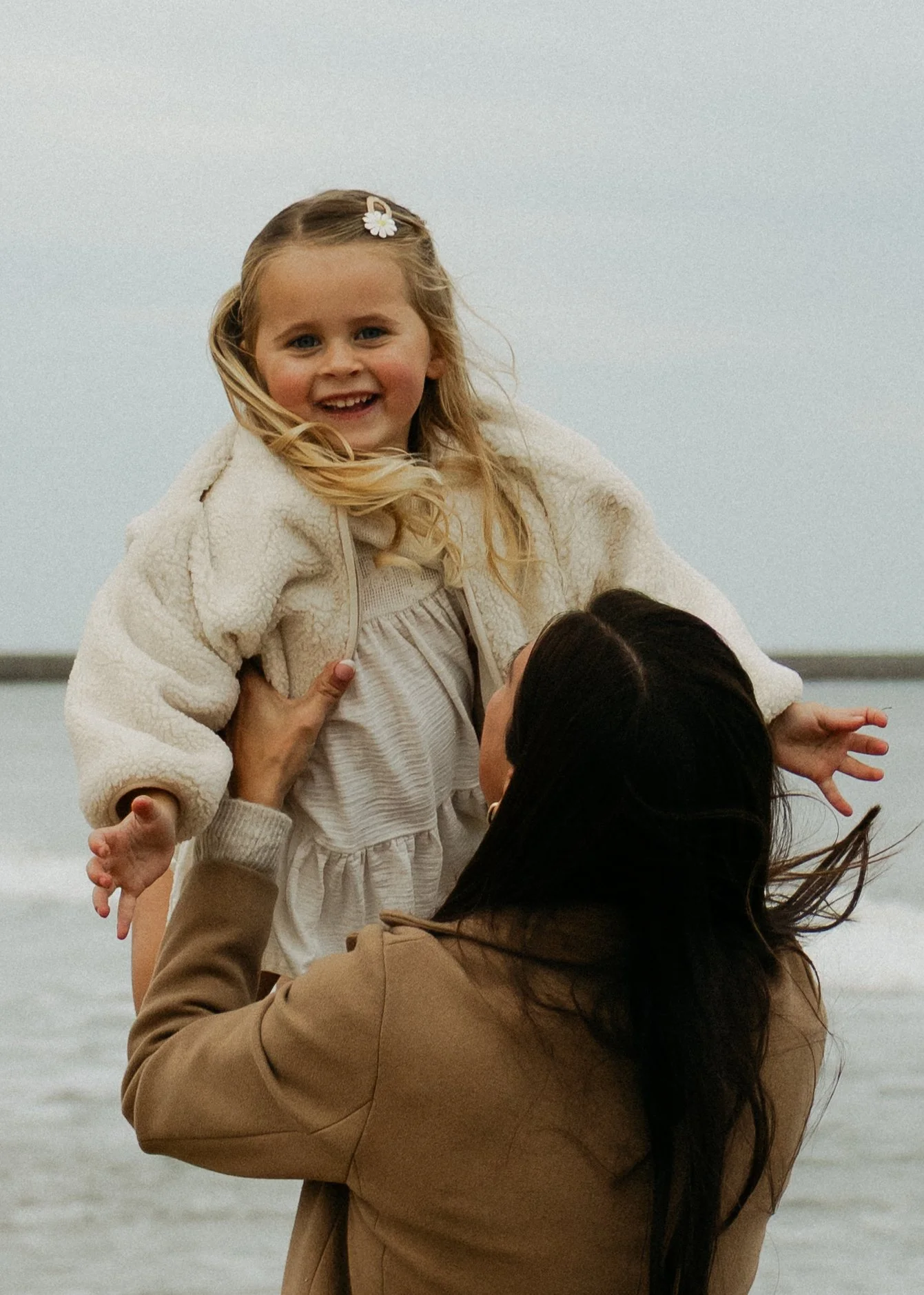 A young girl with blonde hair in a braid, wearing a cream hoodie and fluffy jacket, being held up by an adult woman at the beach, with water and overcast sky in the background.