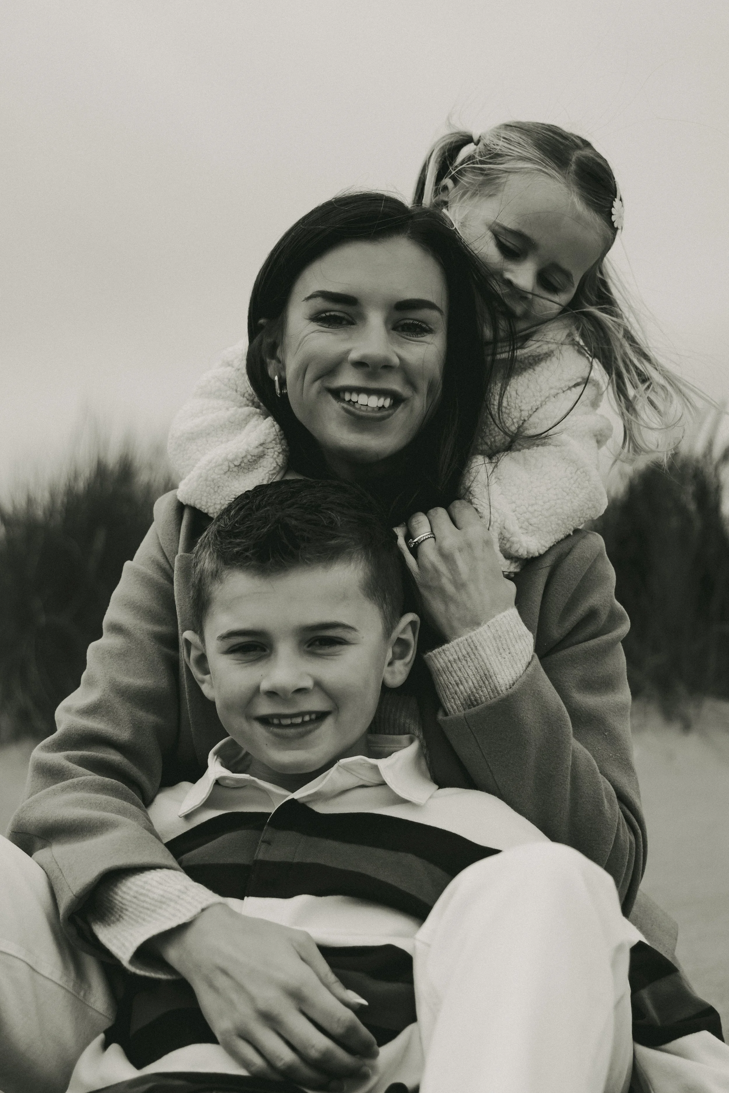 A woman and two children outdoors. The woman has dark hair, smiling with teeth showing, and is wearing a coat with a fleece collar. The girl, with light hair in pigtails and floral hair clips, is on her back leaning to the side, also smiling gently. The boy, with short hair, sitting in front, is smiling with teeth showing, and wearing a striped shirt. They appear happy and close together in a natural setting.