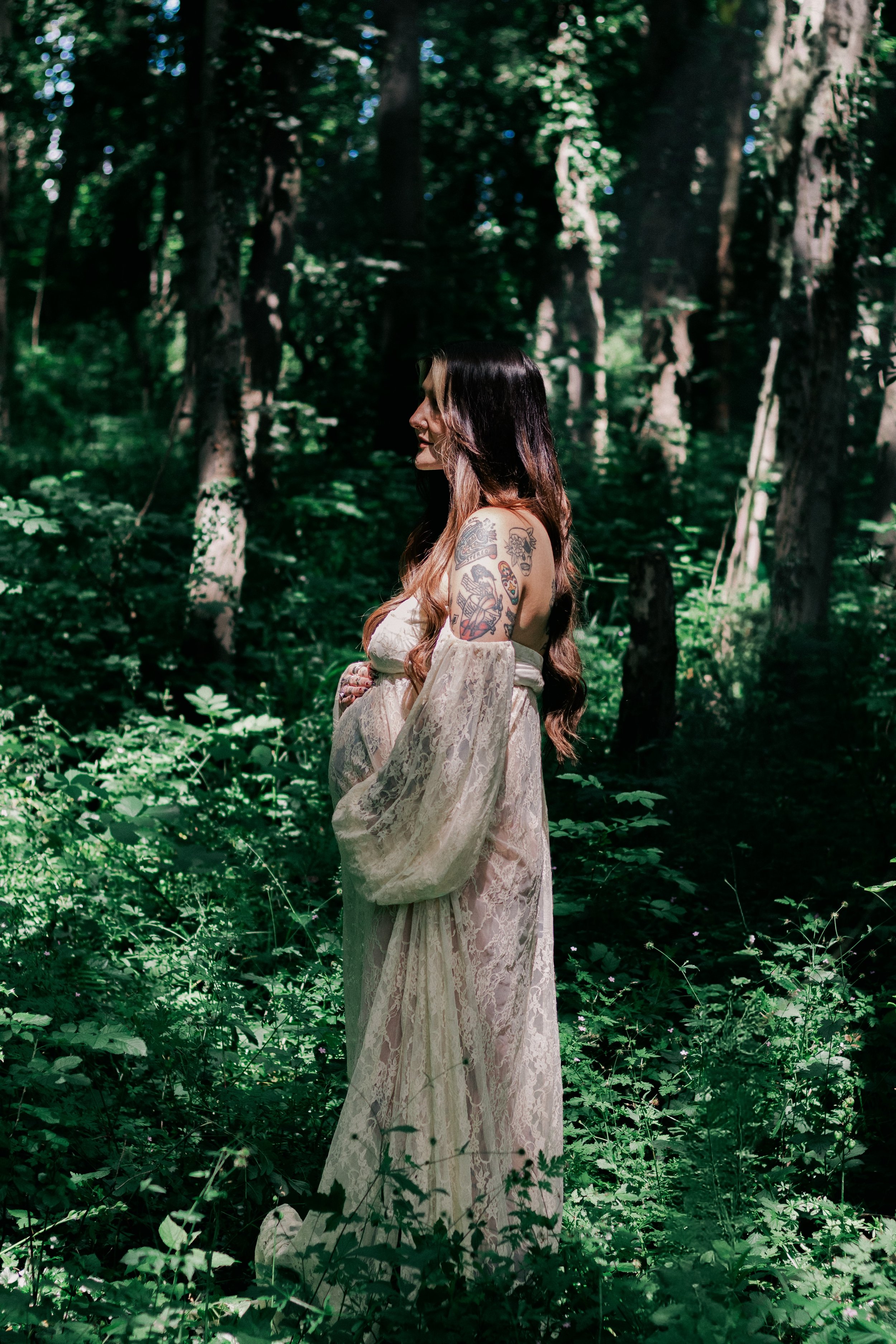 A woman with long wavy hair and tattoos on her arm, wearing a vintage-style white lace dress, standing in a lush green forest with sunlight filtering through the trees.