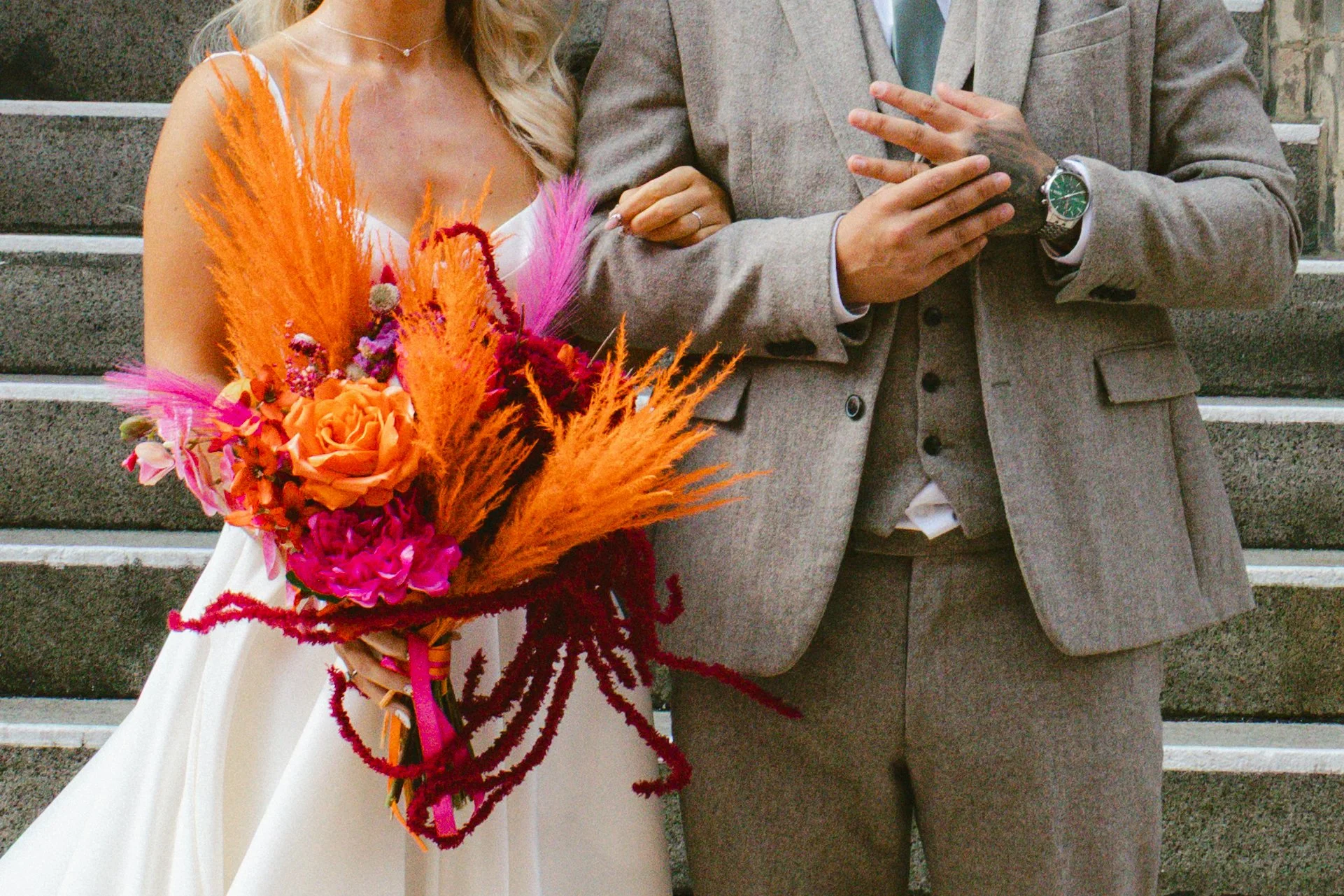 Close-up of a bride holding a colorful bouquet and an escort with a gray suit, both standing on stone steps.