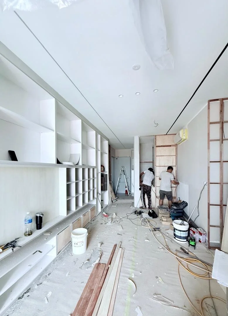 Interior view of a room under construction with two workers installing fixtures or wall panels. Construction tools, materials, and debris are scattered on the floor, with built-in white shelving on the left and an air conditioning unit on the right.