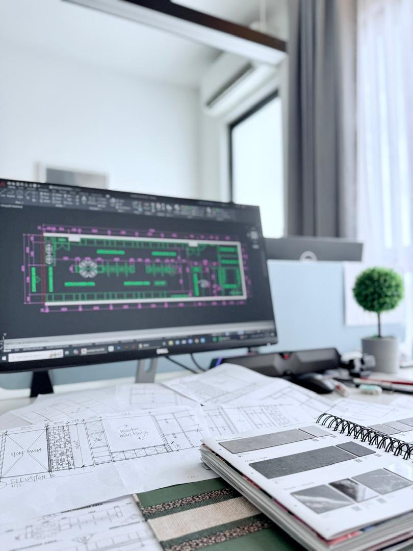 A workspace with architectural blueprints, color sample swatches, a computer monitor displaying a CAD drawing, and a small potted plant on the desk.