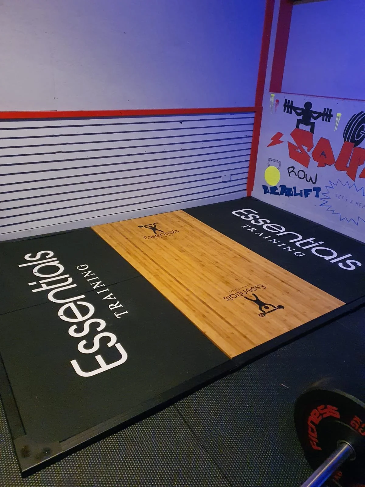 The floor of a gym features branded exercise mats with the words "Essential Strength Training" and "Essensh" and includes a wooden section with the Essensh logo, along with cones and a wall with fitness-related illustrations and text.