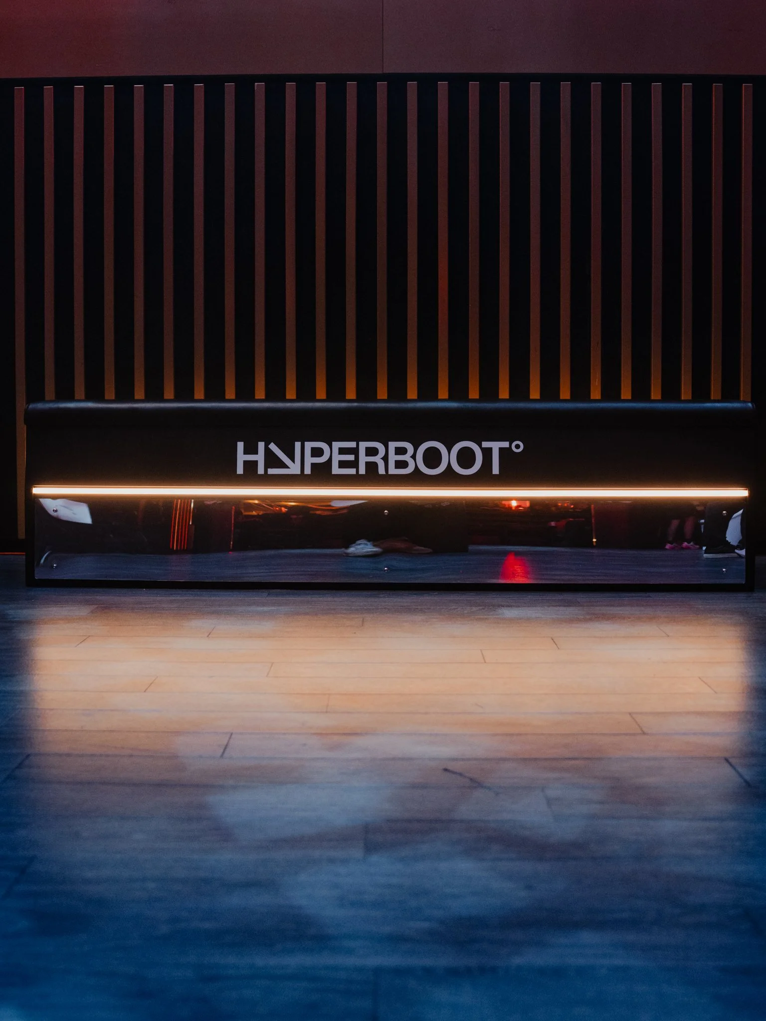 A gym reception desk with a black front panel featuring the white Hyperboot logo and a wooden slat wall behind it, with a light brown wood floor in the foreground.