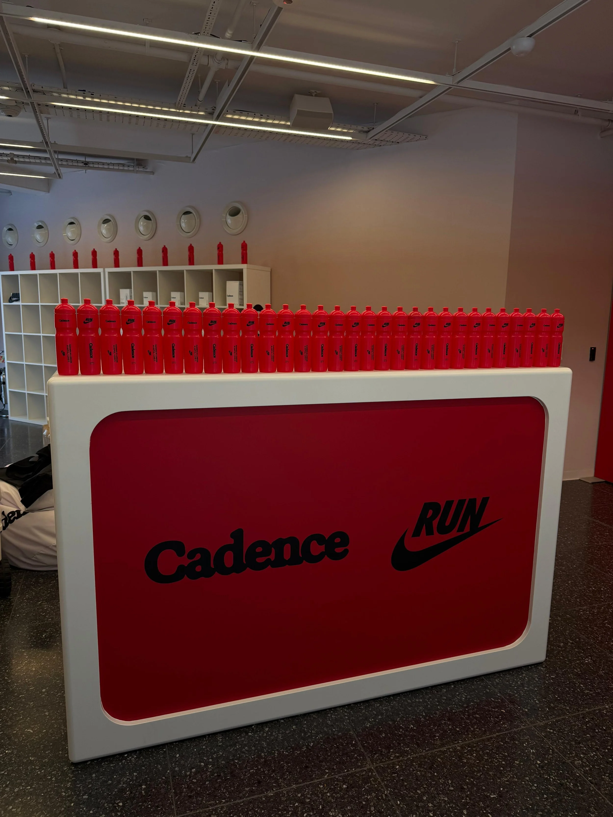 A white table with a red front panel displaying the words 'Cadence' and the Nike swoosh logo, surrounded by multiple red water bottles on top. In the background, there is a white shelving unit and a wall with circular vents, visible in an indoor spac
