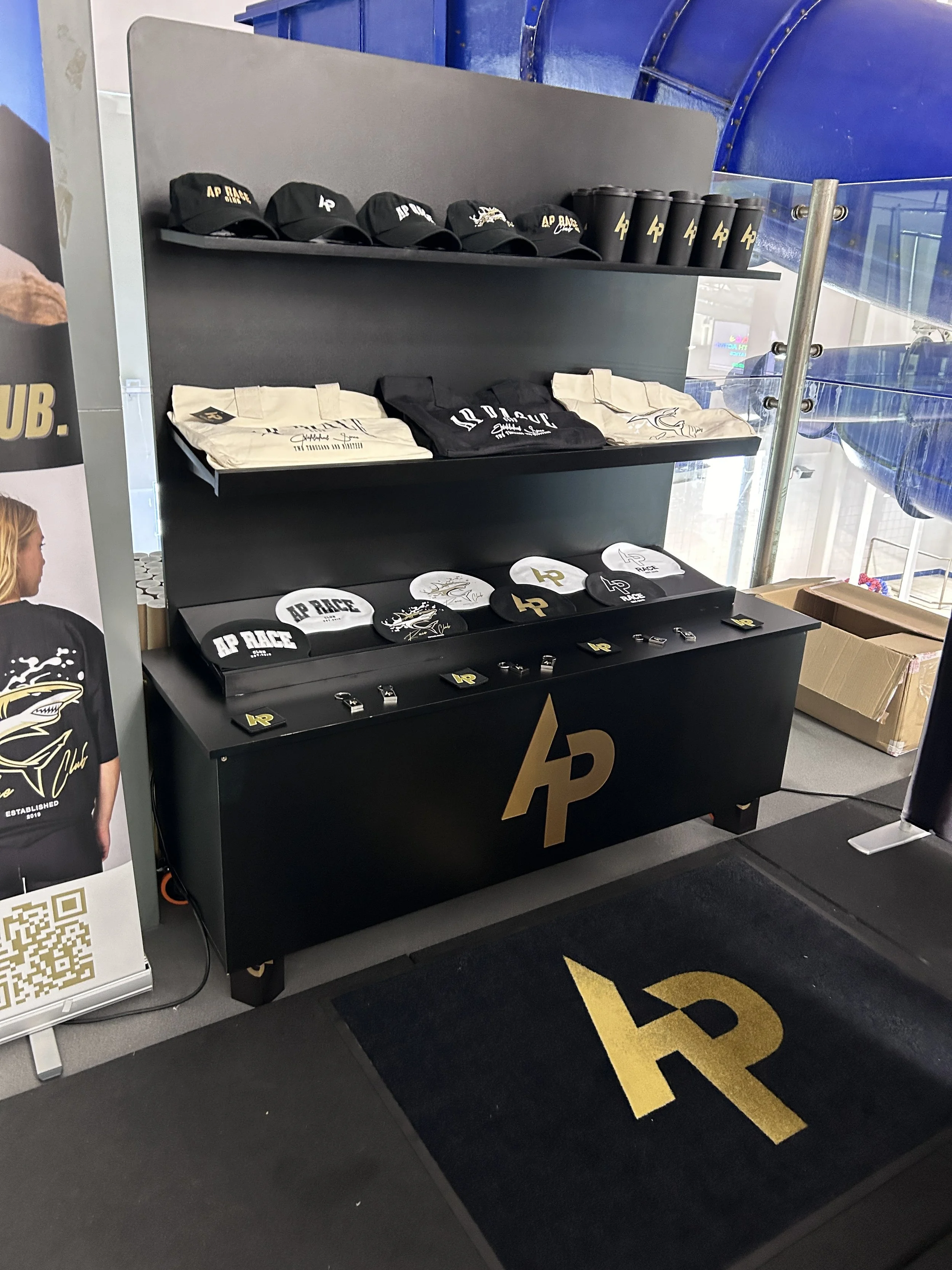 Display shelf with black hats, black tumblers, and white and black caps, all featuring the AP Race logo, in a store or promotional area.