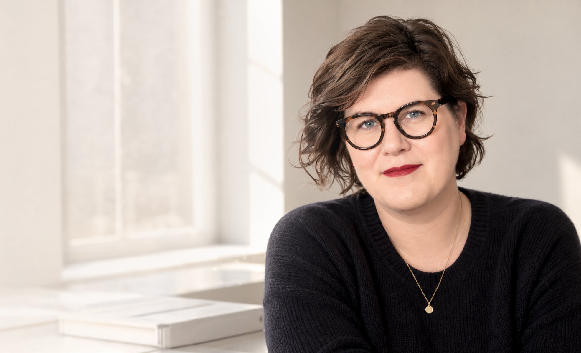 Pia Bennedsen with short wavy brown hair and glasses looks friendly into the camera wearing a black sweater and gold jewelry, sitting near a window.
