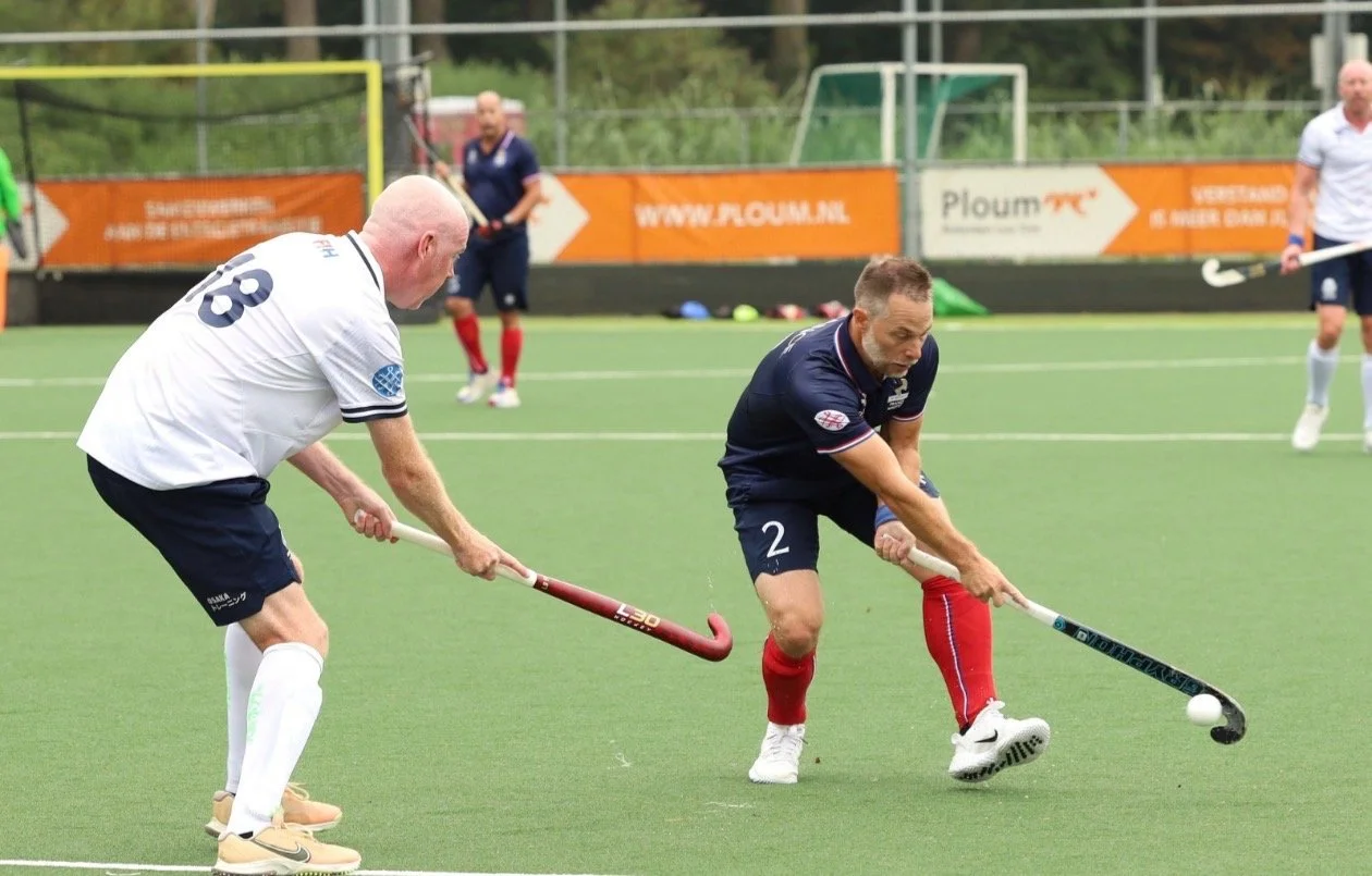 Two men playing field hockey on a green turf. One man in a white shirt and black shorts is leaning forward with a hockey stick, and another man in a dark blue shirt and shorts with red socks is also in a bent posture with a hockey stick, both competing for the ball to gain control.
