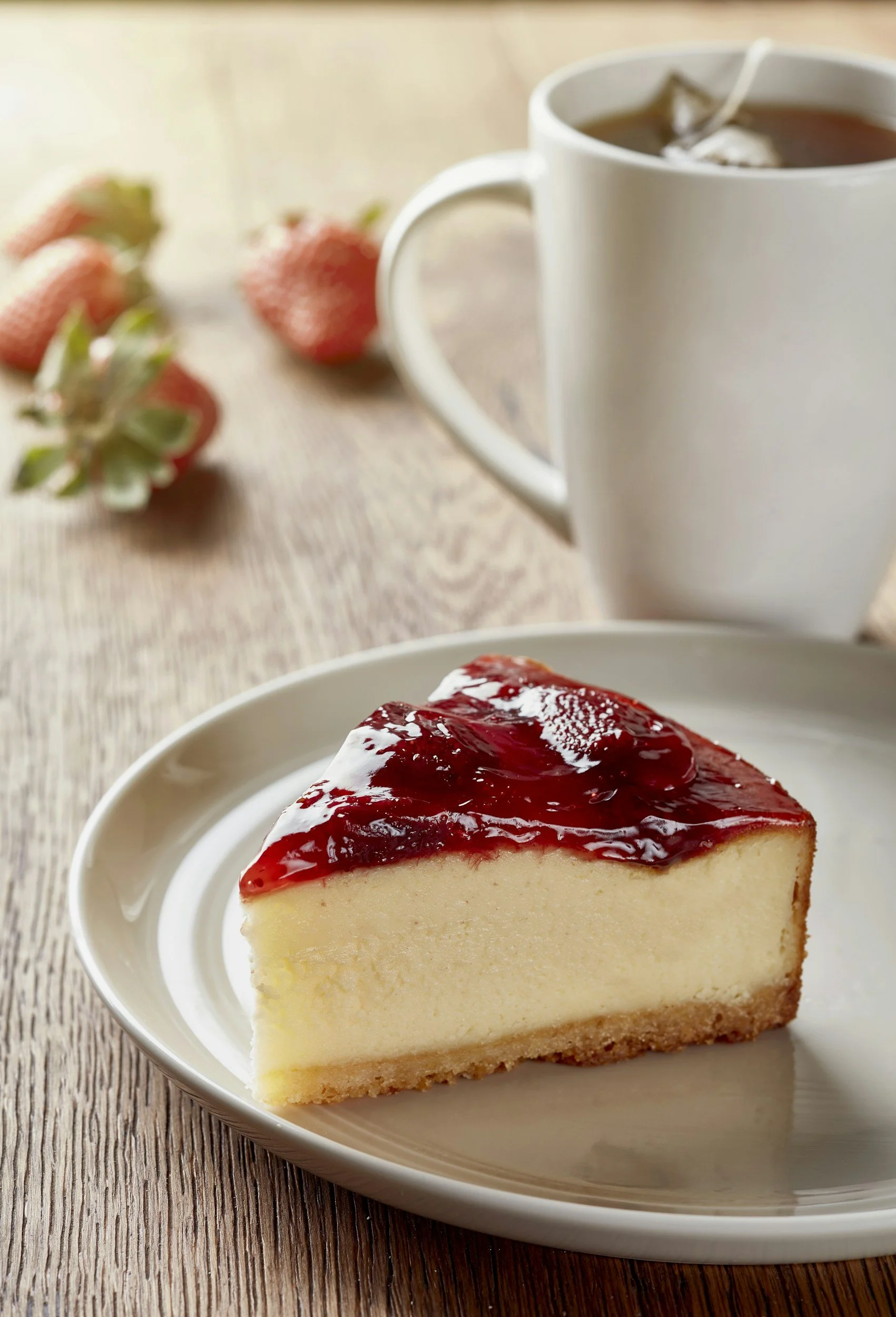 A slice of cheesecake with a graham cracker crust and topped with strawberry glaze on a white plate. In the background, a white mug of tea with a teabag and a few strawberries on a wooden table.