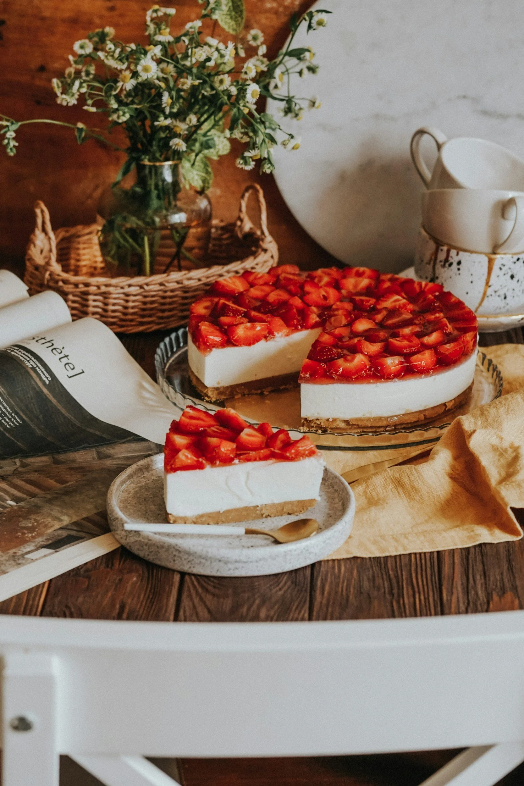 A slice of strawberry cheesecake on a small gray plate, with whole strawberries on top, placed on a wooden table. In the background, there is a jar of daisies, a white teapot, cups, and a large white bowl.