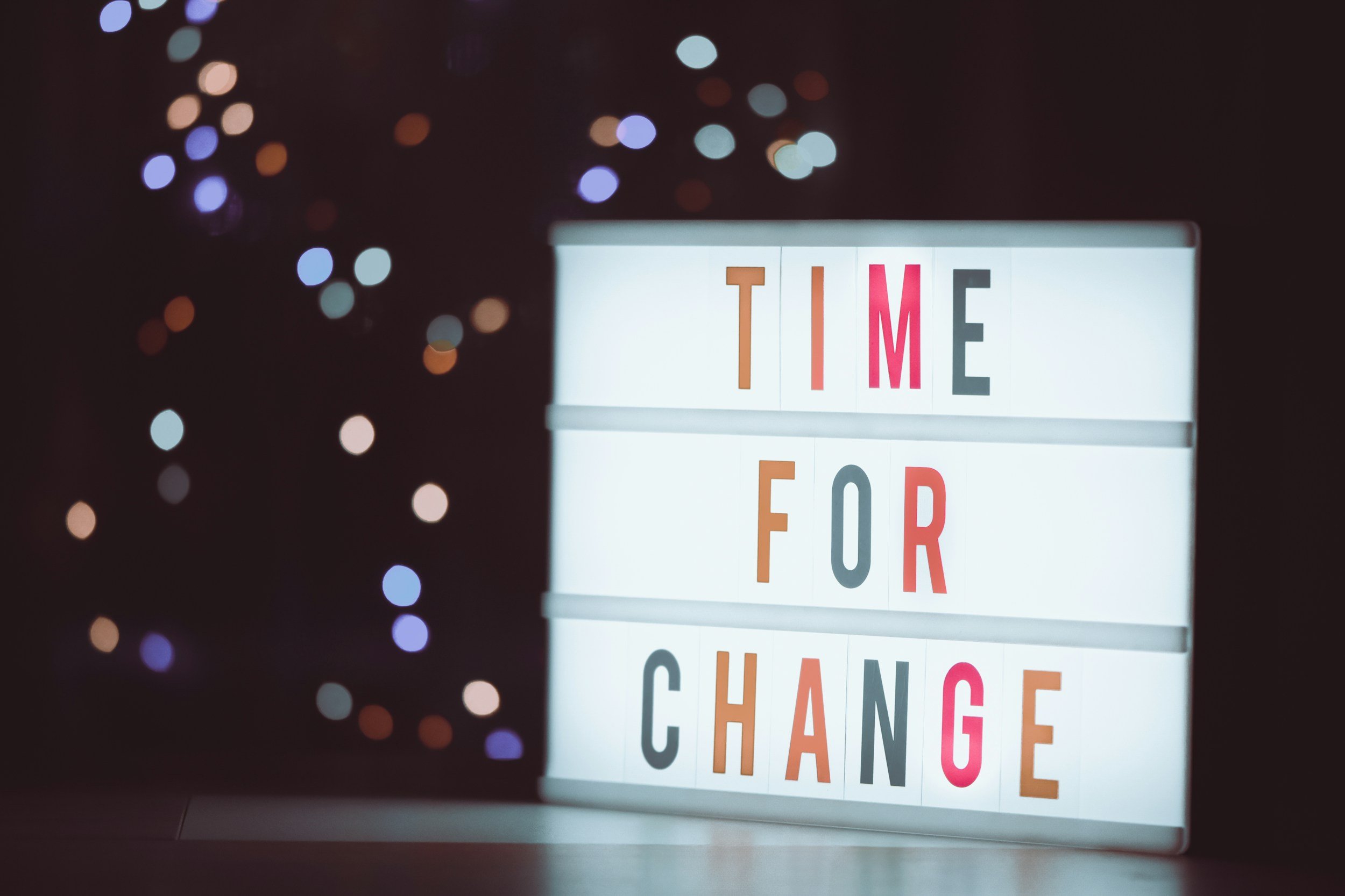 Lightbox sign with the words 'TIME FOR CHANGE' in colorful letters, in front of a dark background with blurred colorful lights.