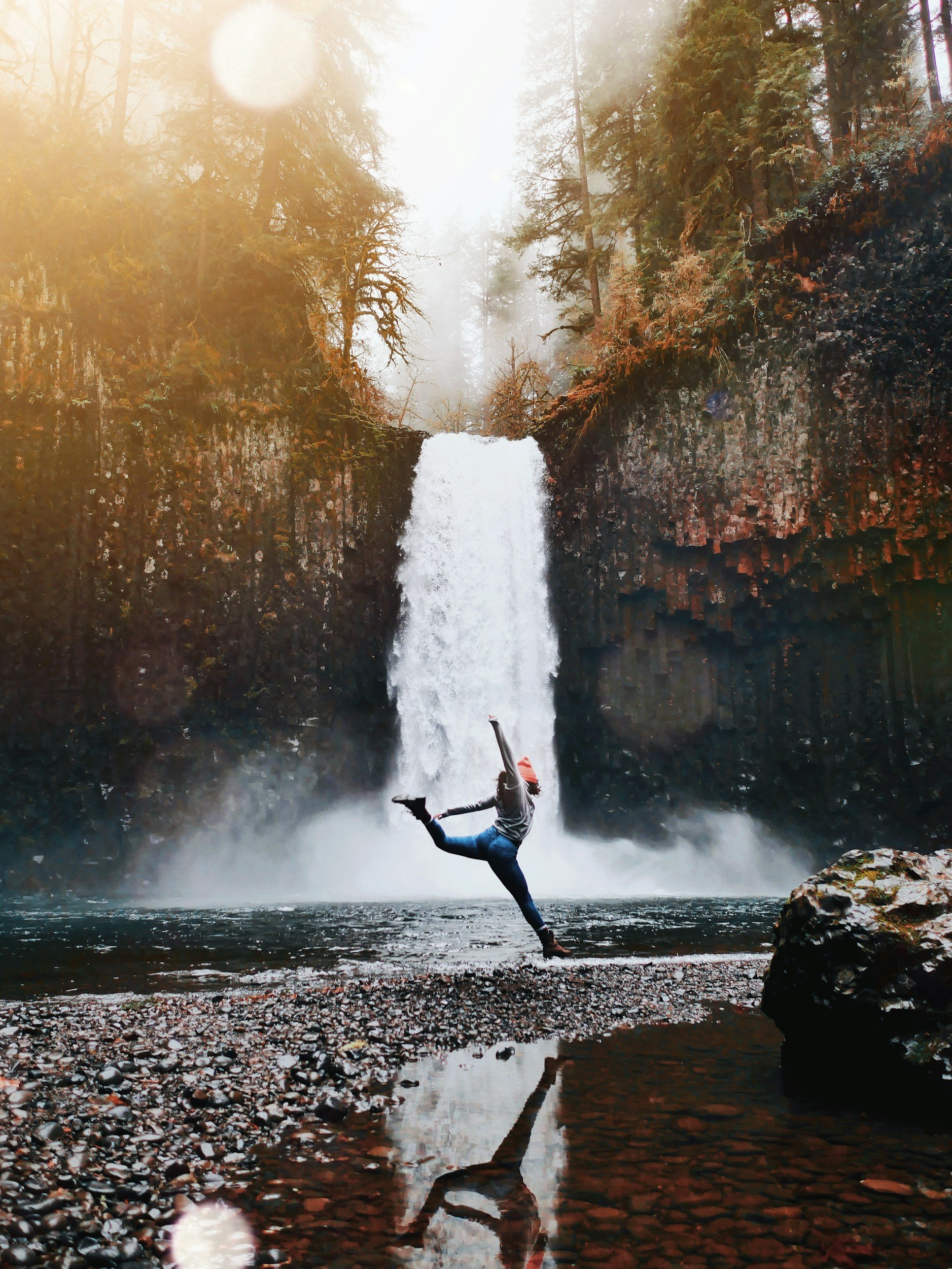 Free and empowering image of a woman balancing on one leg in front of a waterfall in a forest with mist and sunlight