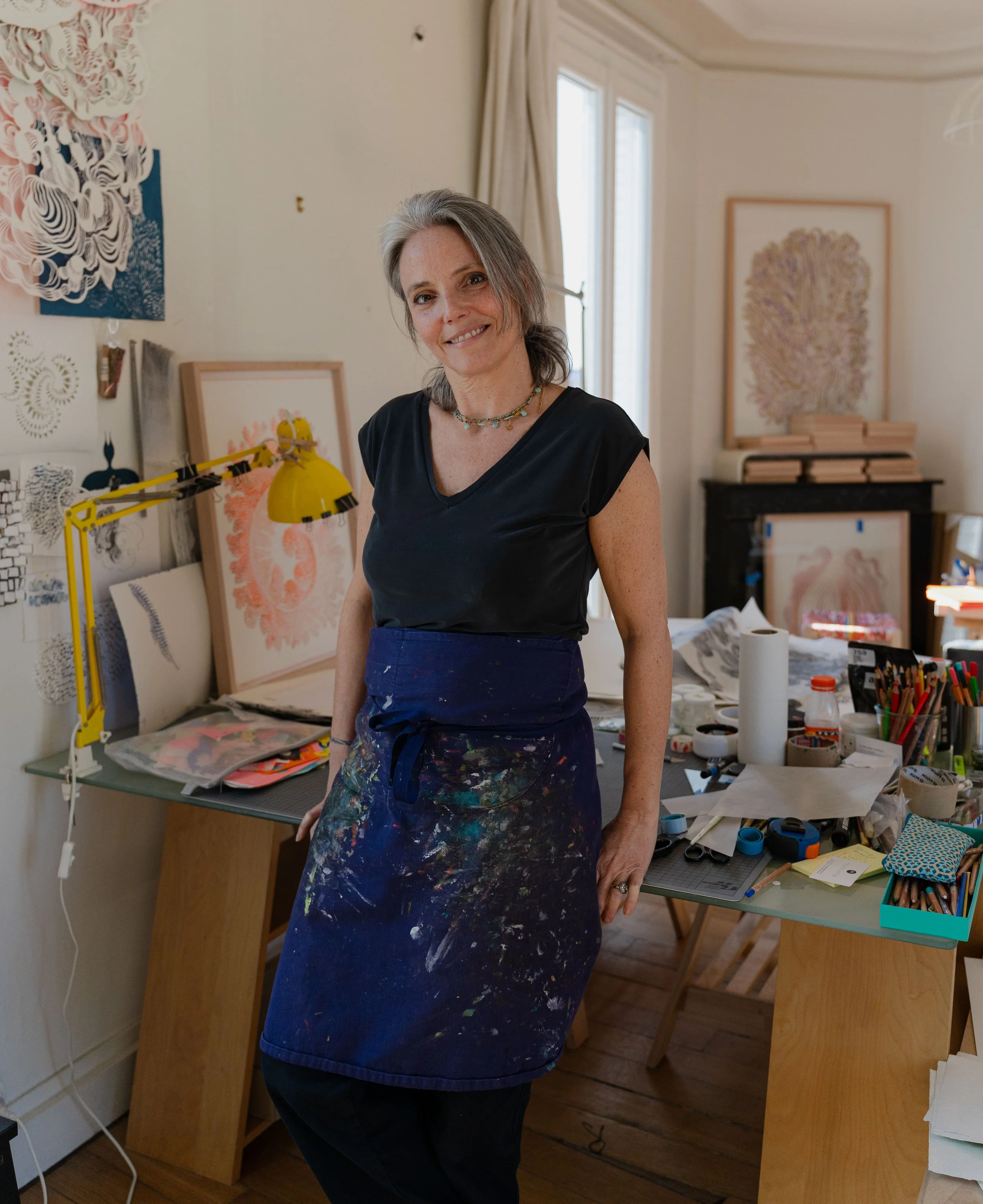 Une femme souriante avec des cheveux gris, portant un haut noir et une jupe avec des taches de peinture, dans un atelier d'art ou un studio, entourée de projets artistiques, de fournitures d'art et de peintures en arrière-plan.