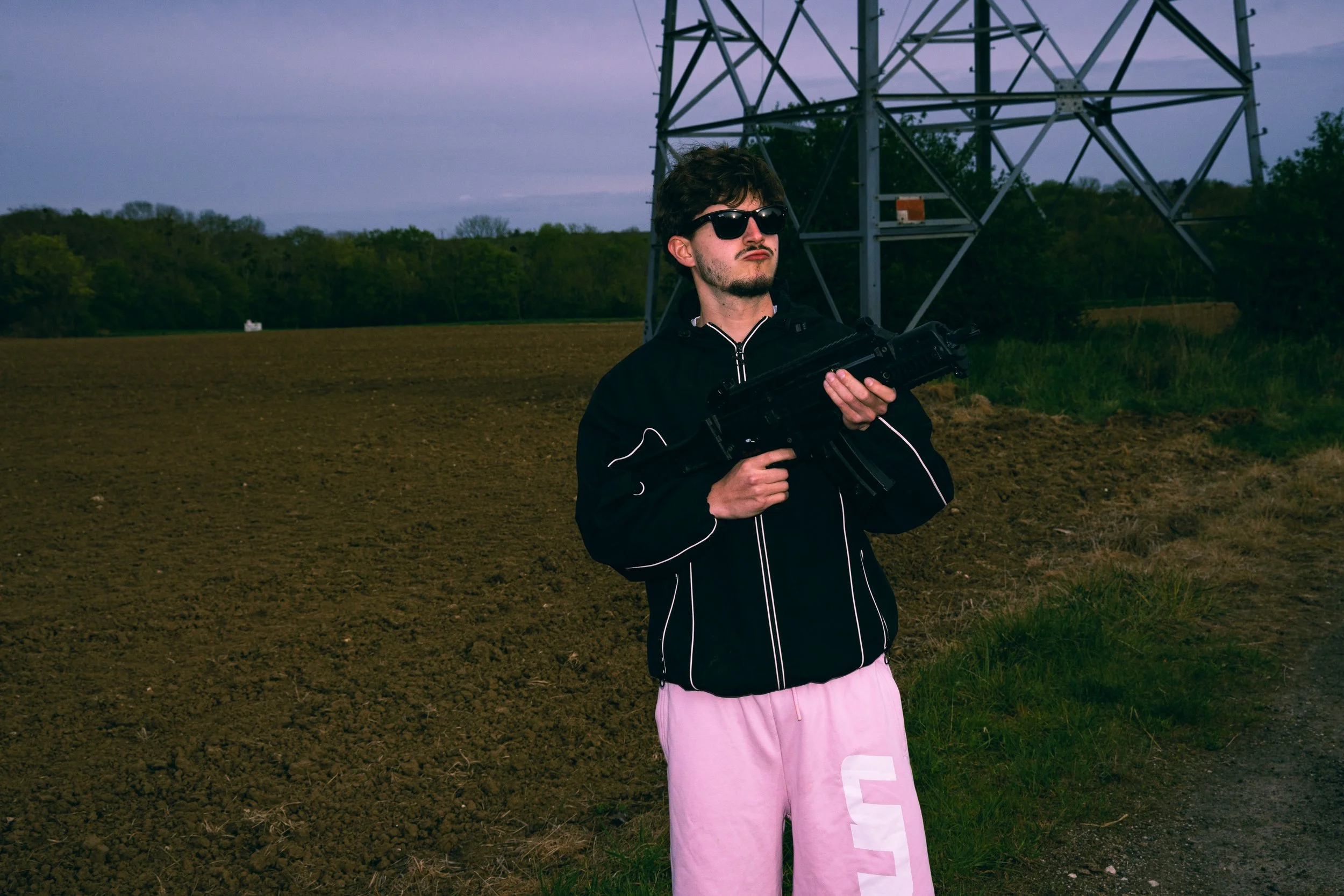 Jeune homme portant des lunettes de soleil, un sweat noir, et un pantalon rose clair avec une lettre blanche, tenant un fusil, devant une tour électrique dans un champ en pleine nature, au crépuscule.