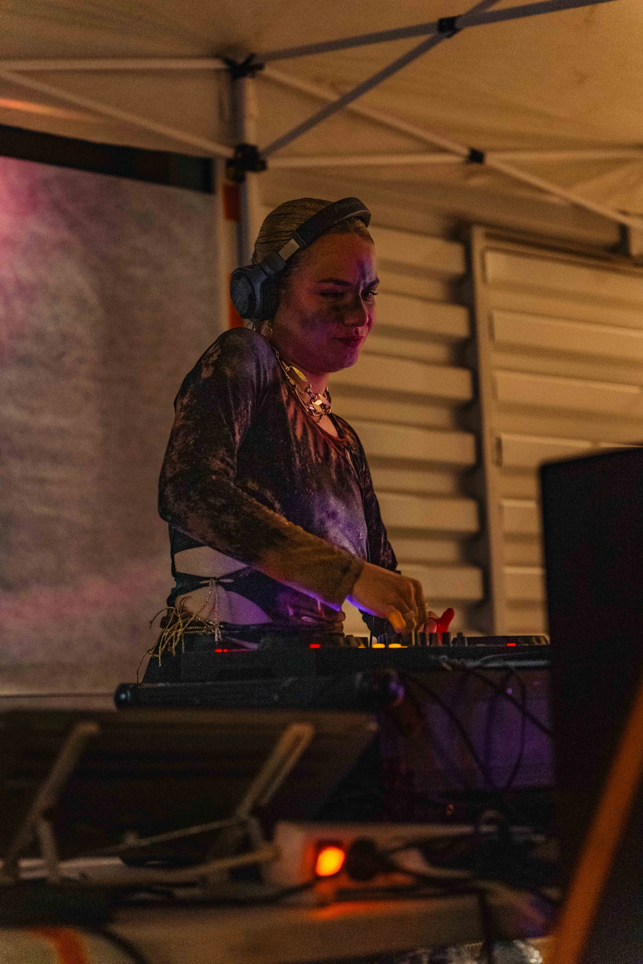 Une femme DJ avec des cheveux blonds, portant un casque de DJ et une chaîne en or, utilisant une table de mixage dans un environnement sombre avec un éclairage tamisé.
