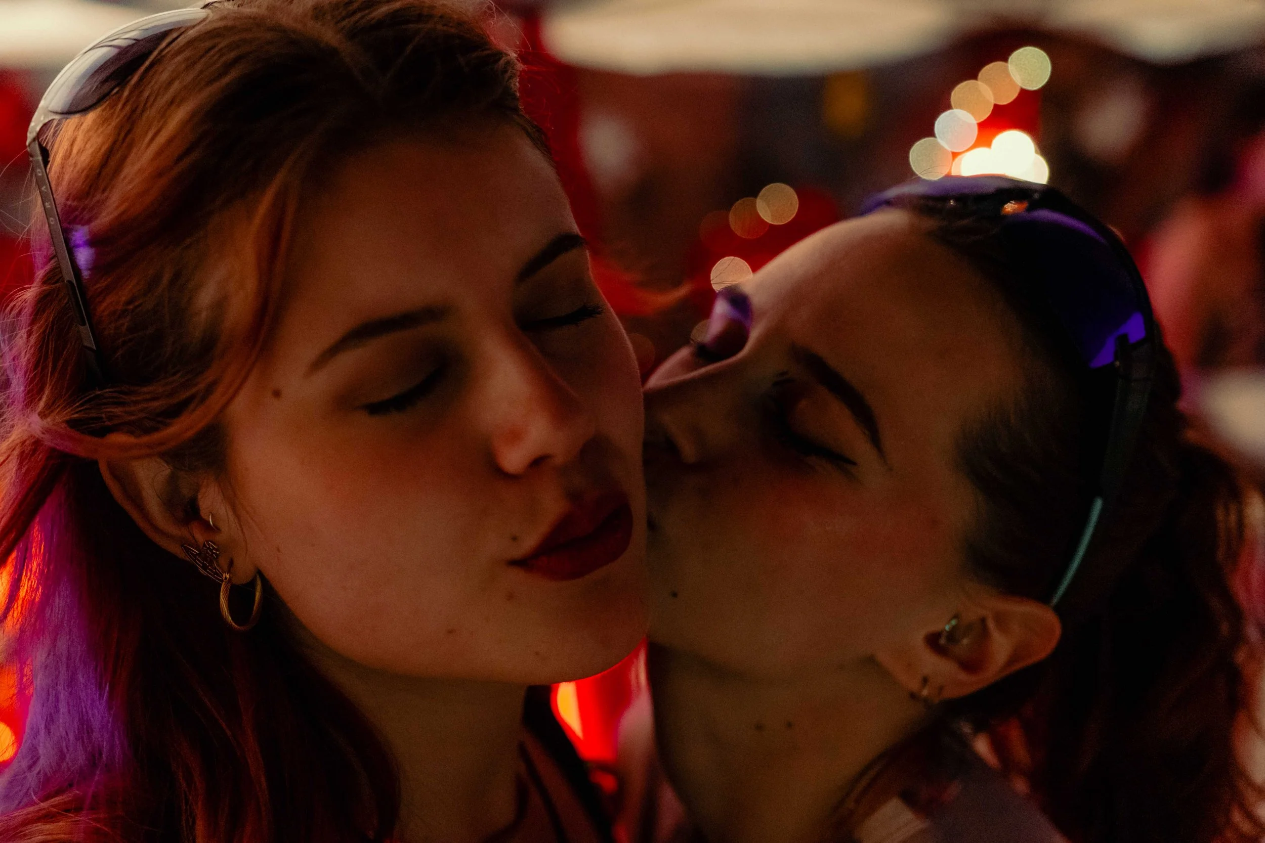 Deux personnes très proches, dans un environnement sombre avec des lumières colorées bokeh en arrière-plan. La femme a des cheveux roux et porte des accessoires, l'homme porte des lunettes avec des lumières LED. Ils semblent partager un moment intime