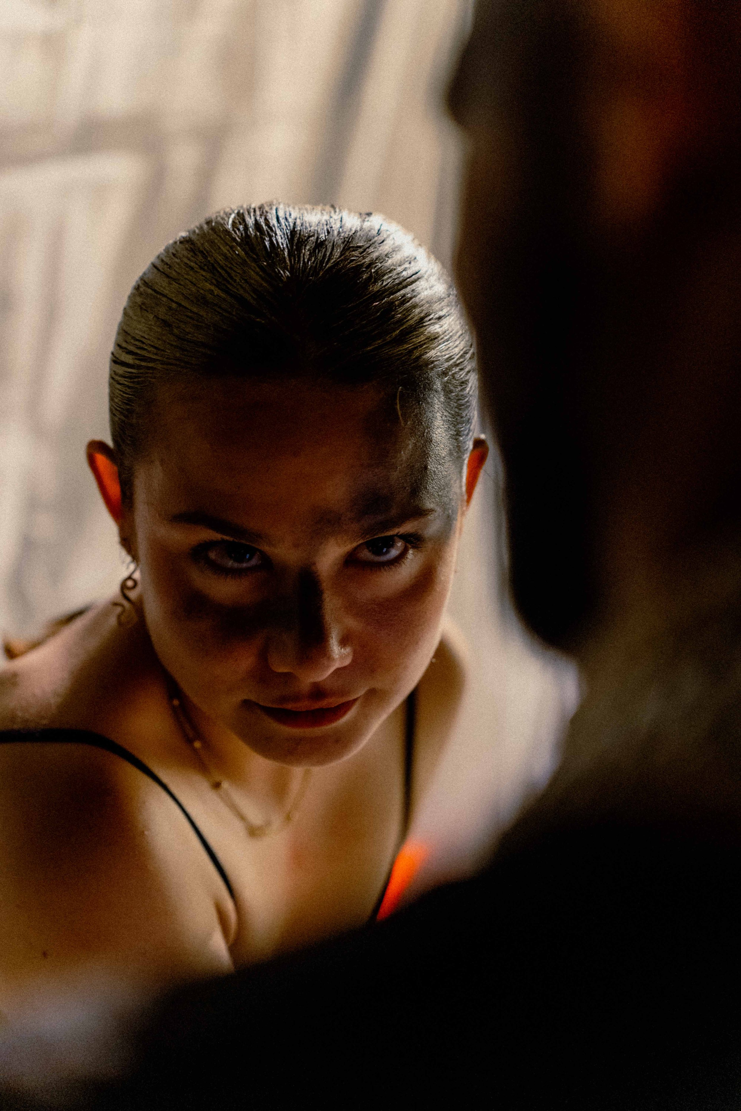 Une femme avec des cheveux coiffés en chignon, portant un haut noir, regardant directement l'objectif, avec un éclairage chaud et une ambiance intime.