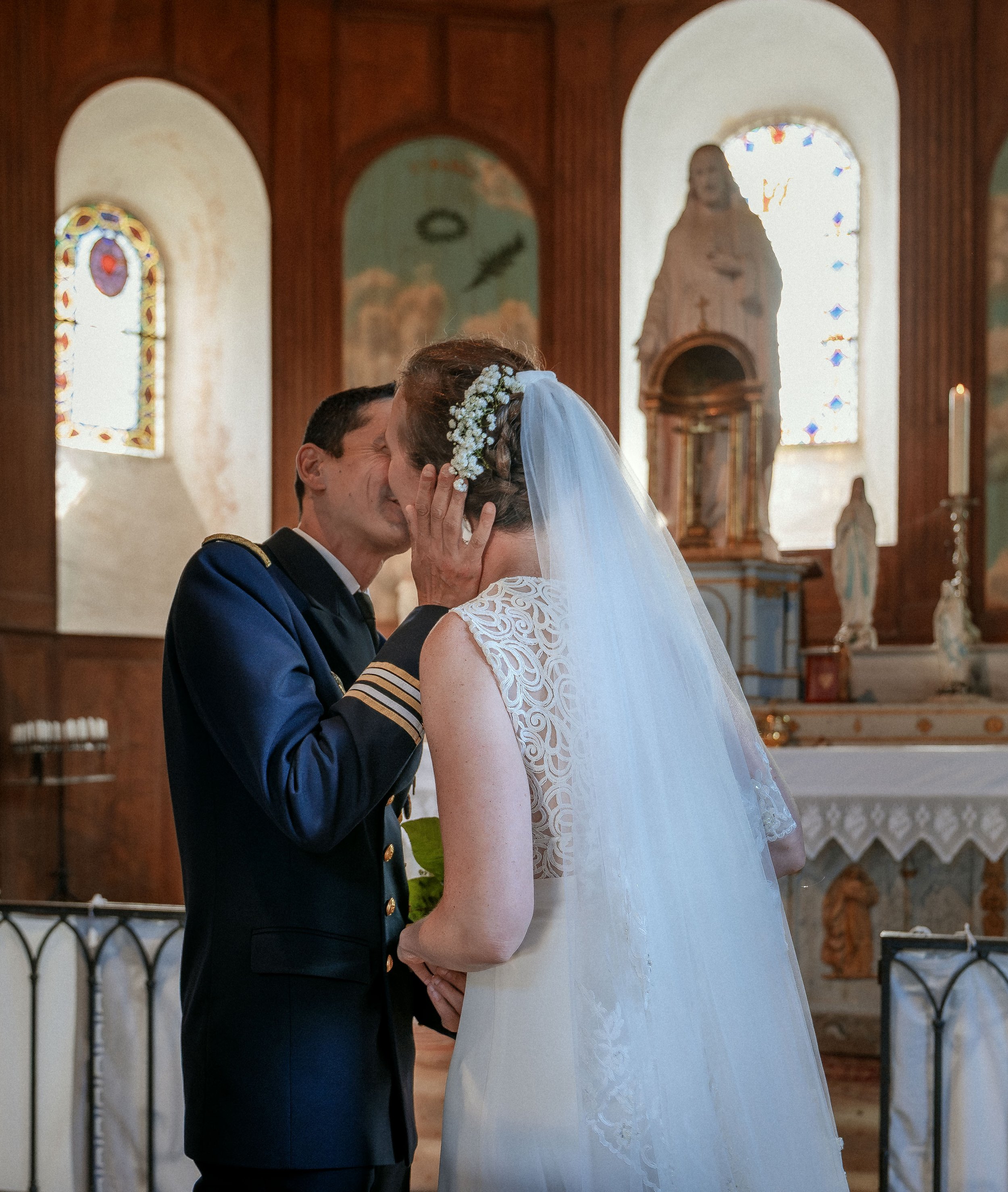 Un couple de mariés échange un baiser dans une église, la femme porte une robe de mariée blanche avec un voile et un serre-tête floral, l'homme porte un uniforme militaire.