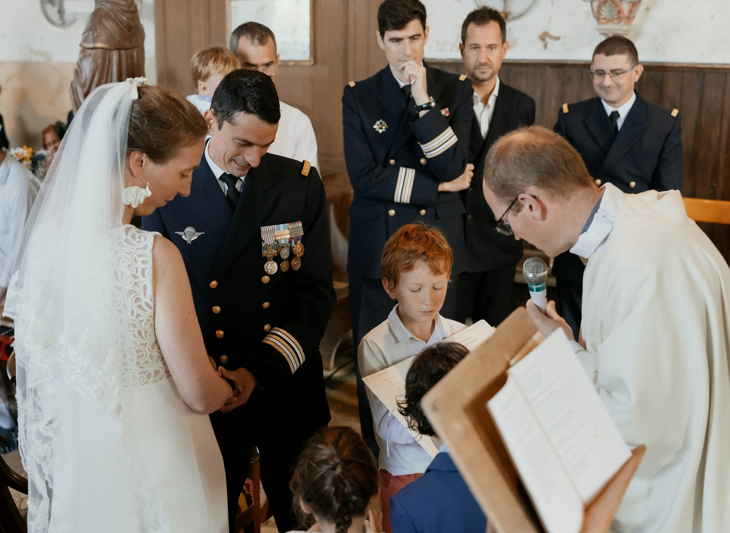 Une cérémonie de mariage civil se déroule dans une église, où une femme en robe de mariée et un homme en uniforme militaire échangent des vœux avec un prêtre. Plusieurs personnes regardent la scène, dont des enfants, certains en uniforme. L'atmosphèr